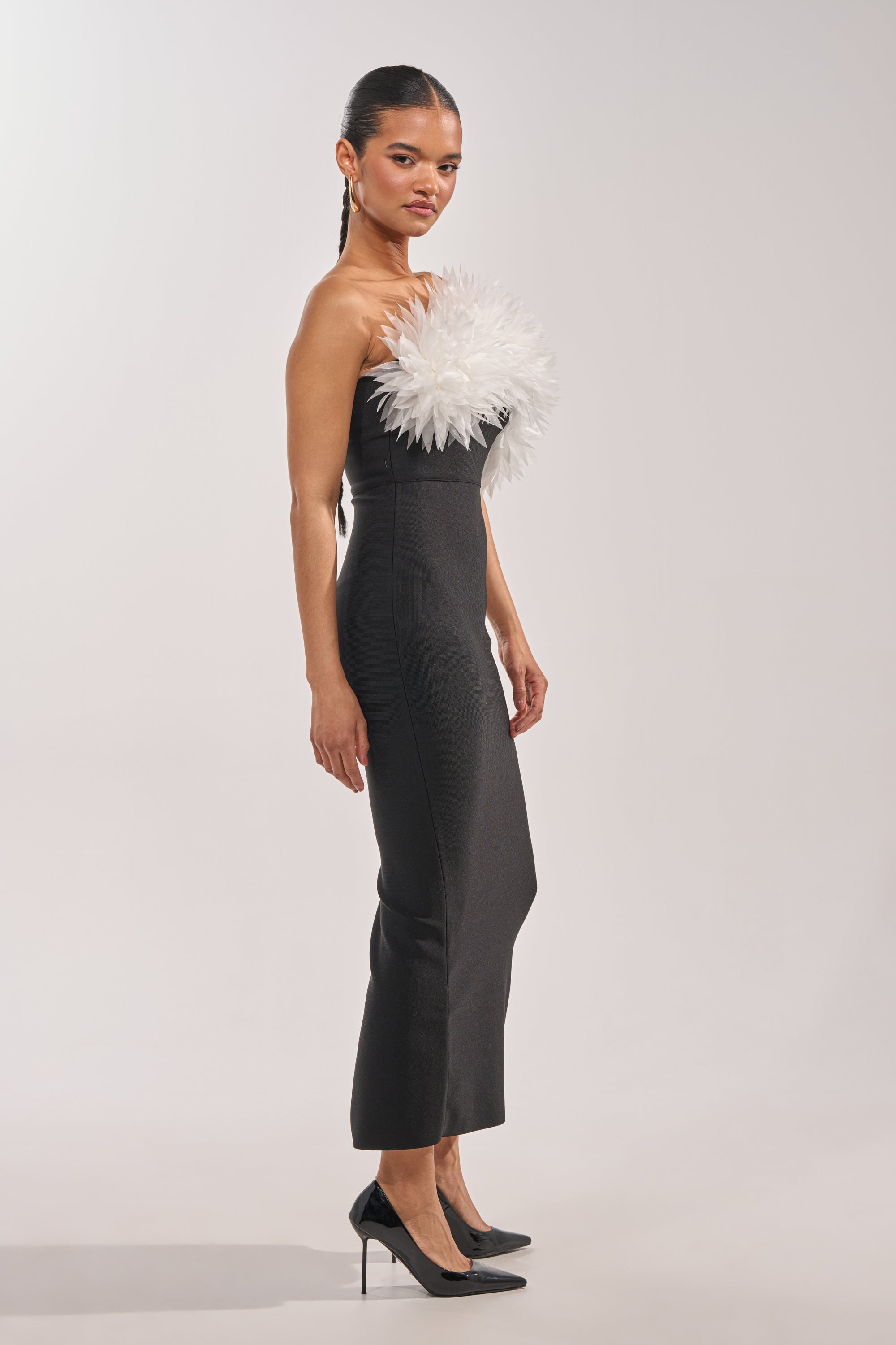 Woman stands in profile wearing the HEART ON MY SLEEVE BANDAGE MIDI DRESS IN BLACK—strapless with bold white feather detail—paired with black heels and sleek hair, embodying modern evening style against a plain light gray background. 2