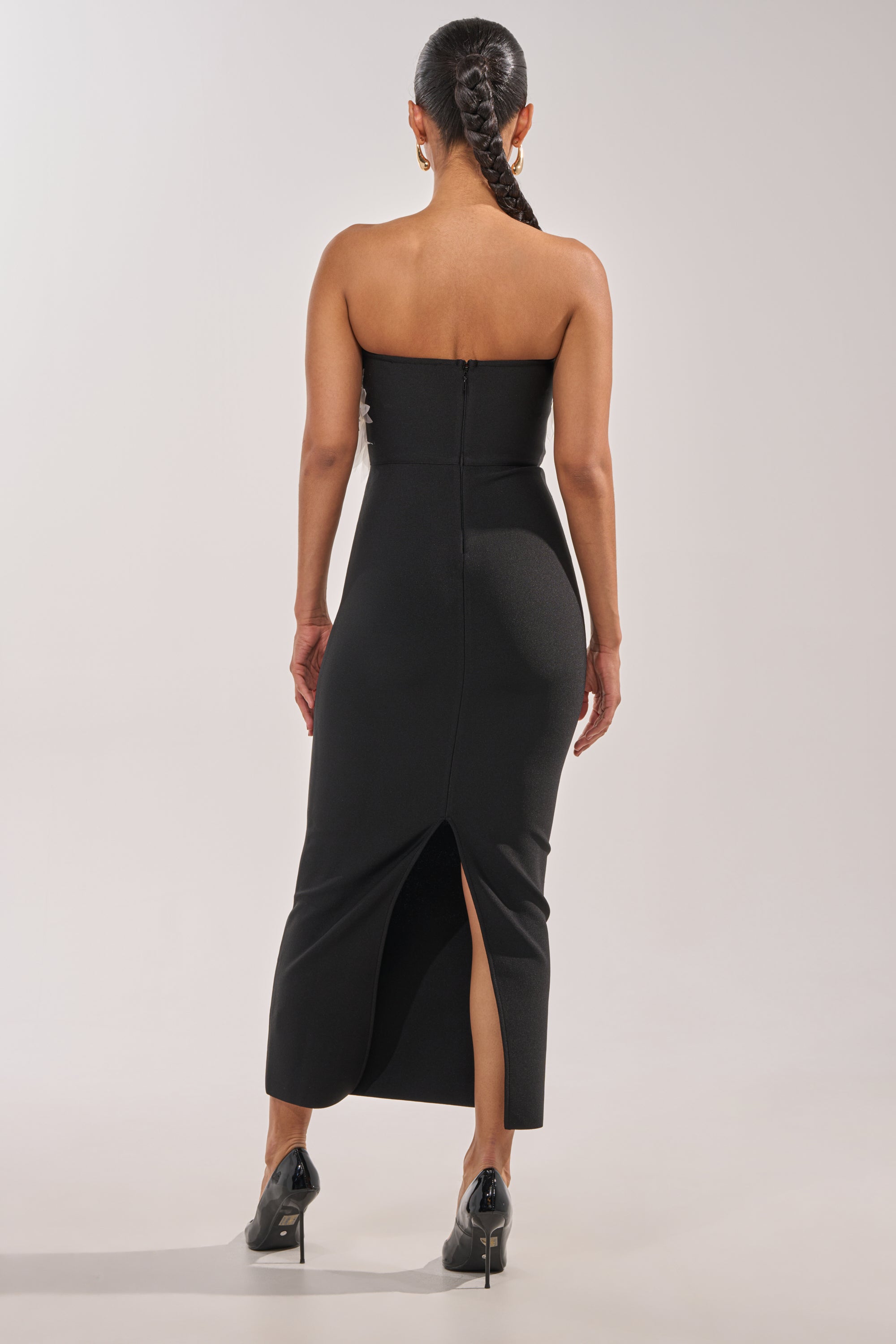 A woman with a long braid wears the HEART ON MY SLEEVE BANDAGE MIDI DRESS IN BLACK—an elegant, strapless, form-fitting midi dress with a back slit—paired with black high heels, standing against a plain light background. 3