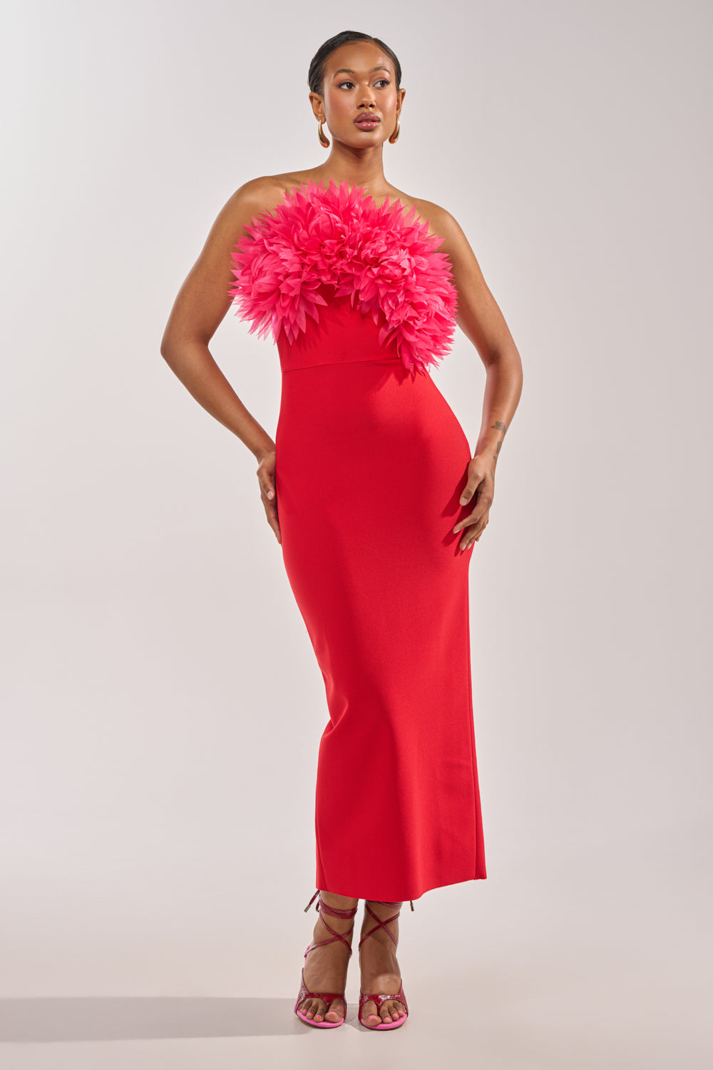 A woman poses in the HEART ON MY SLEEVE Bandage Midi Dress—a strapless red dress with pink feather and 3D flower details—styled with matching heels and hoop earrings, against a plain background, ideal for a wedding guest look.