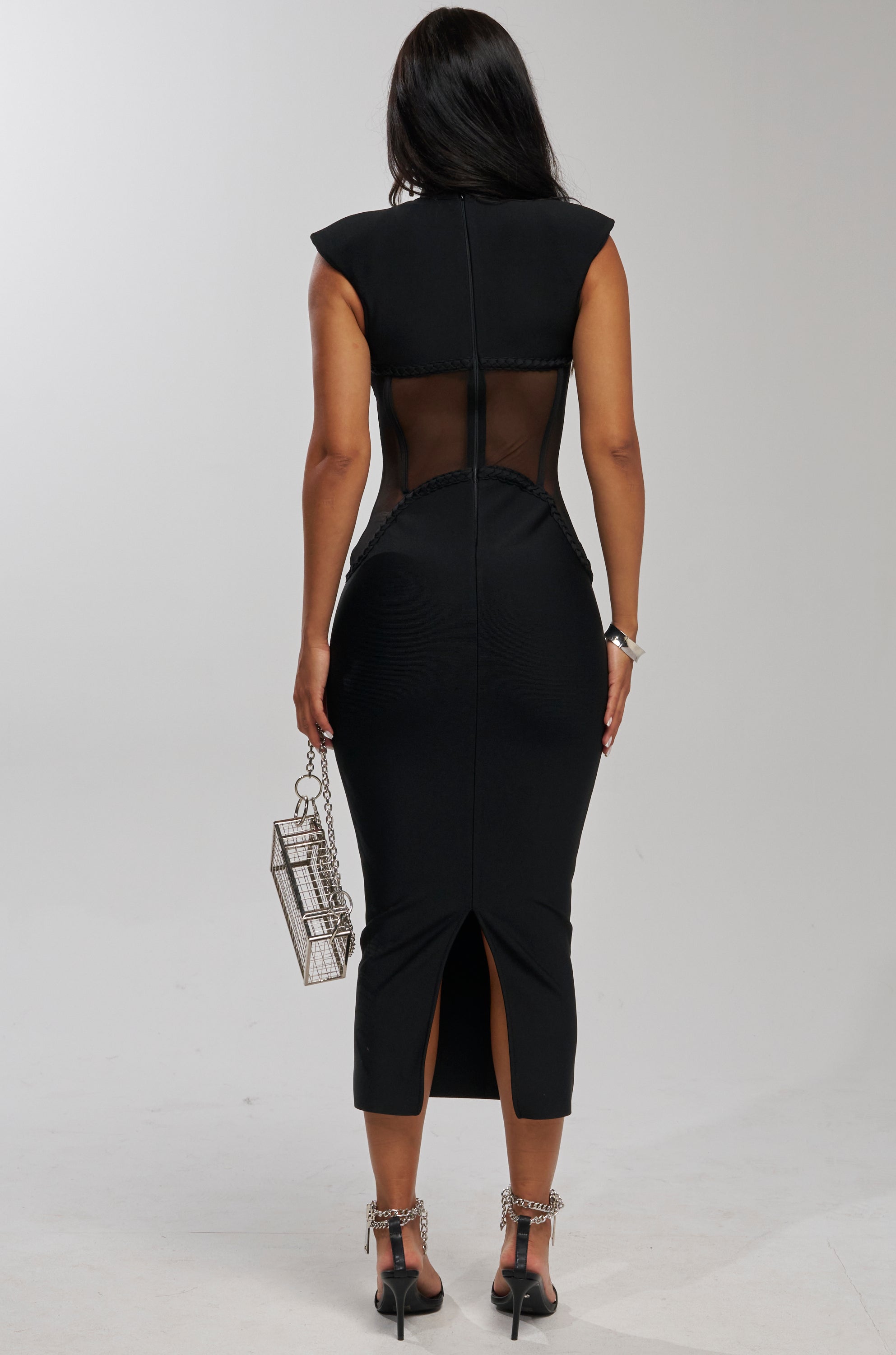 A woman faces away, wearing the ICONIC INSTINCT BANDAGE CORSET MIDI DRESS with sheer back panels, black chain-detailed heels, and holding a small silver boxy purse with a chain strap. The background is plain and light-colored.