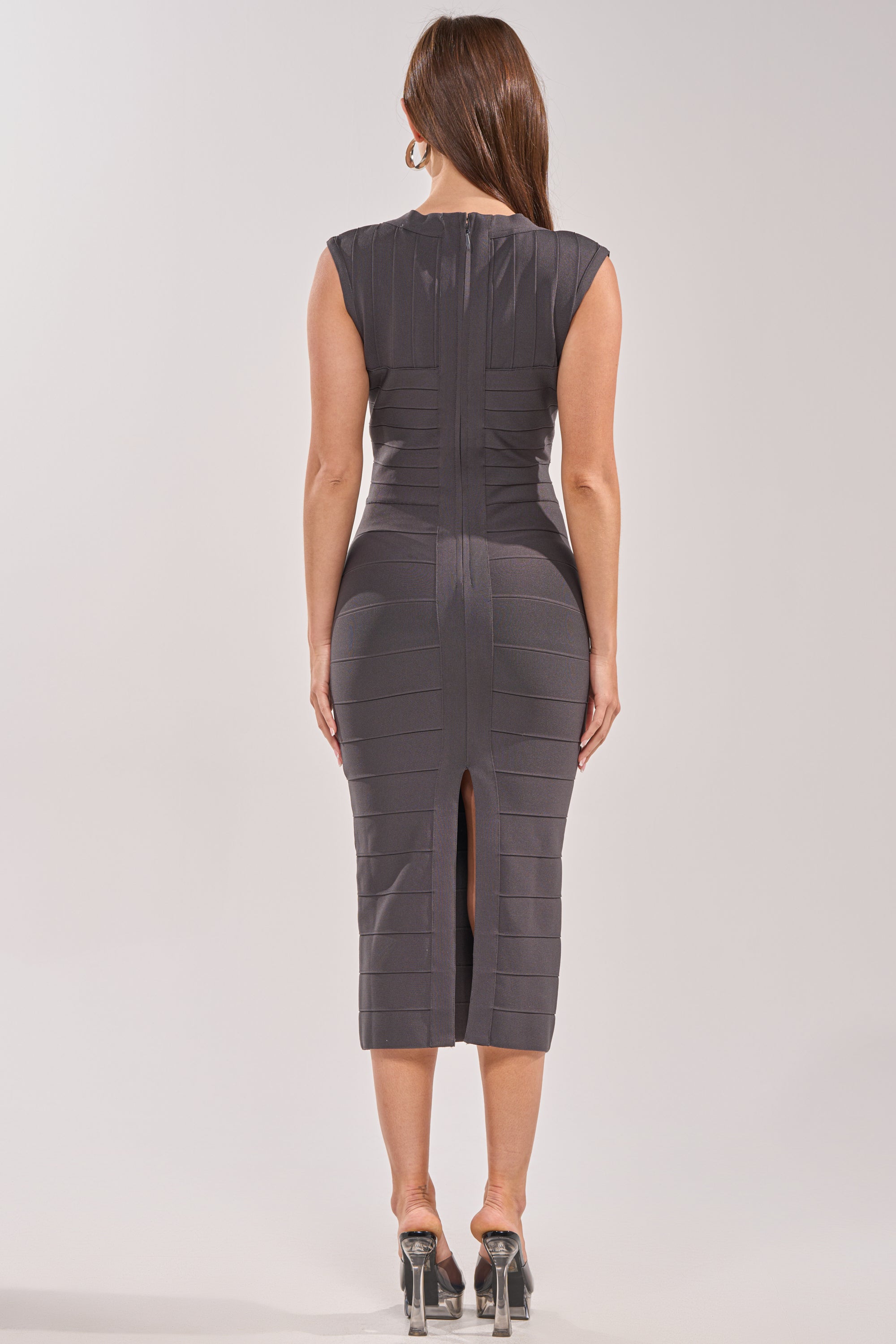 A woman wearing the SOFT GLAM BANDAGE MIDI DRESS—fitted, sleeveless, grey with a back slit—stands with her back to the camera. She has long brown hair and wears high-heeled open-toe shoes, perfect for cocktail parties.