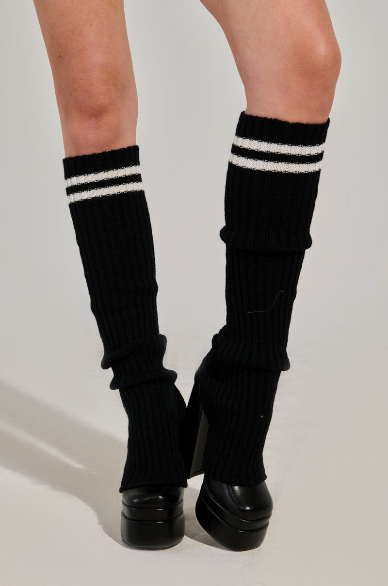 A person wearing TOO TRENDY KNIT LEGWARMERS, featuring a black ribbed knit design with athletic stripes at the top and paired with black platform shoes. The legwarmers have a slouchy silhouette, slightly scrunched, against a plain white background.