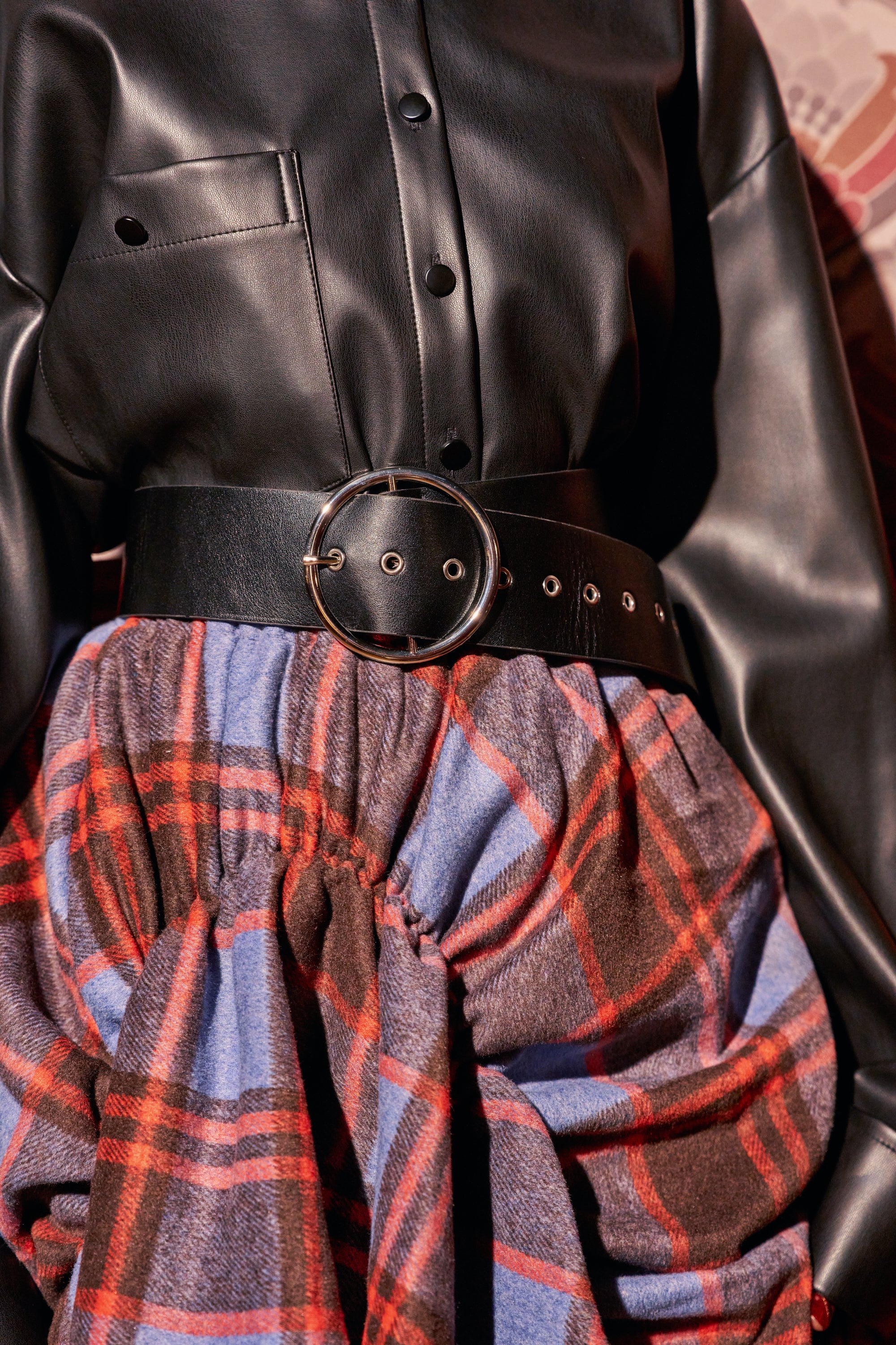 BUCKLED UP BELT, a black faux leather waist belt with a large circular silver buckle worn over a black shirt and a blue and red plaid skirt.