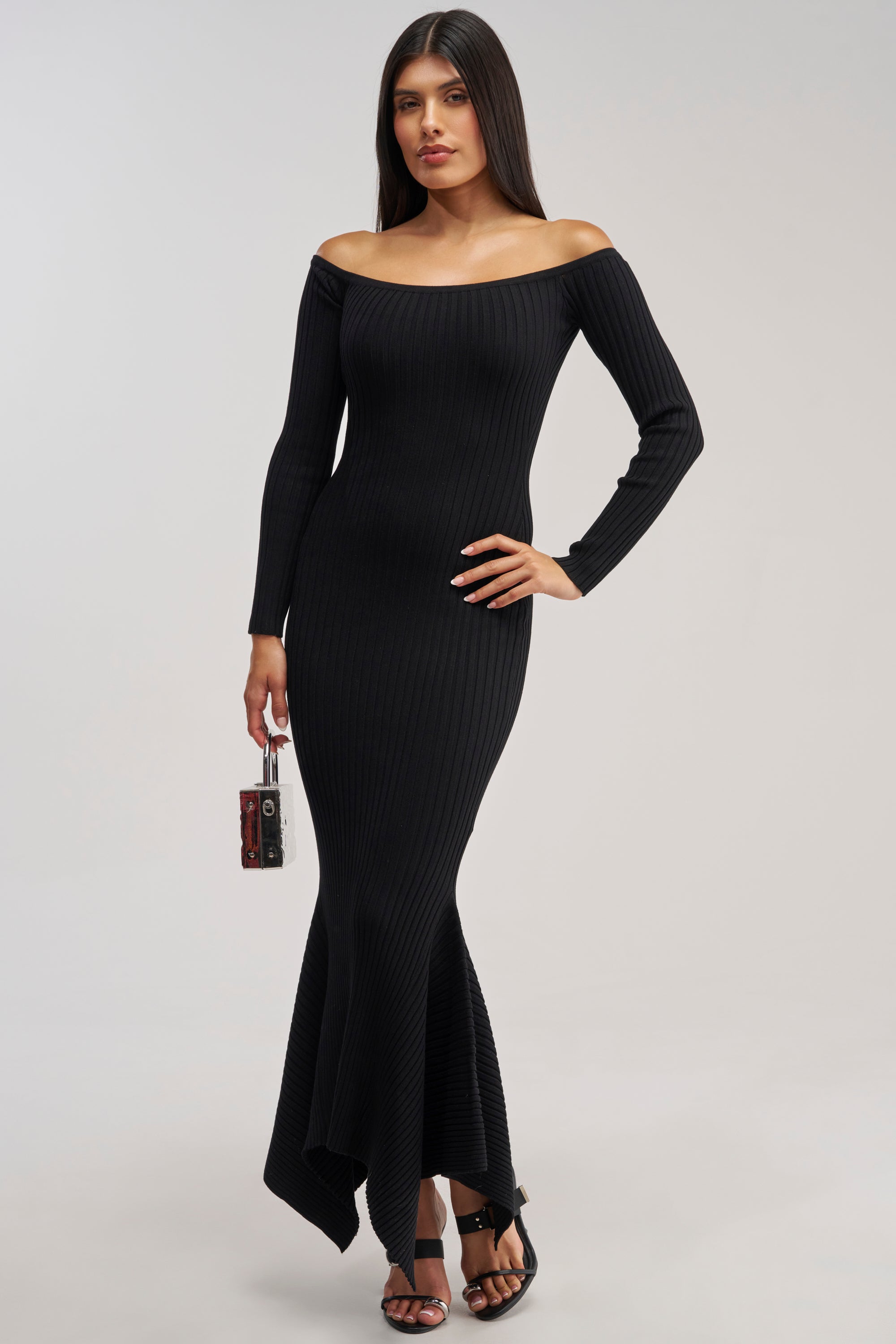 Dressed in the SO SWEET KNIT MAXI DRESS IN BLACK—a long, ribbed, off-the-shoulder sweater dress with elegant long sleeves—she holds a sleek box clutch and wears classic black heels. This chic winter date night look radiates confidence as she stands poised against a light background.