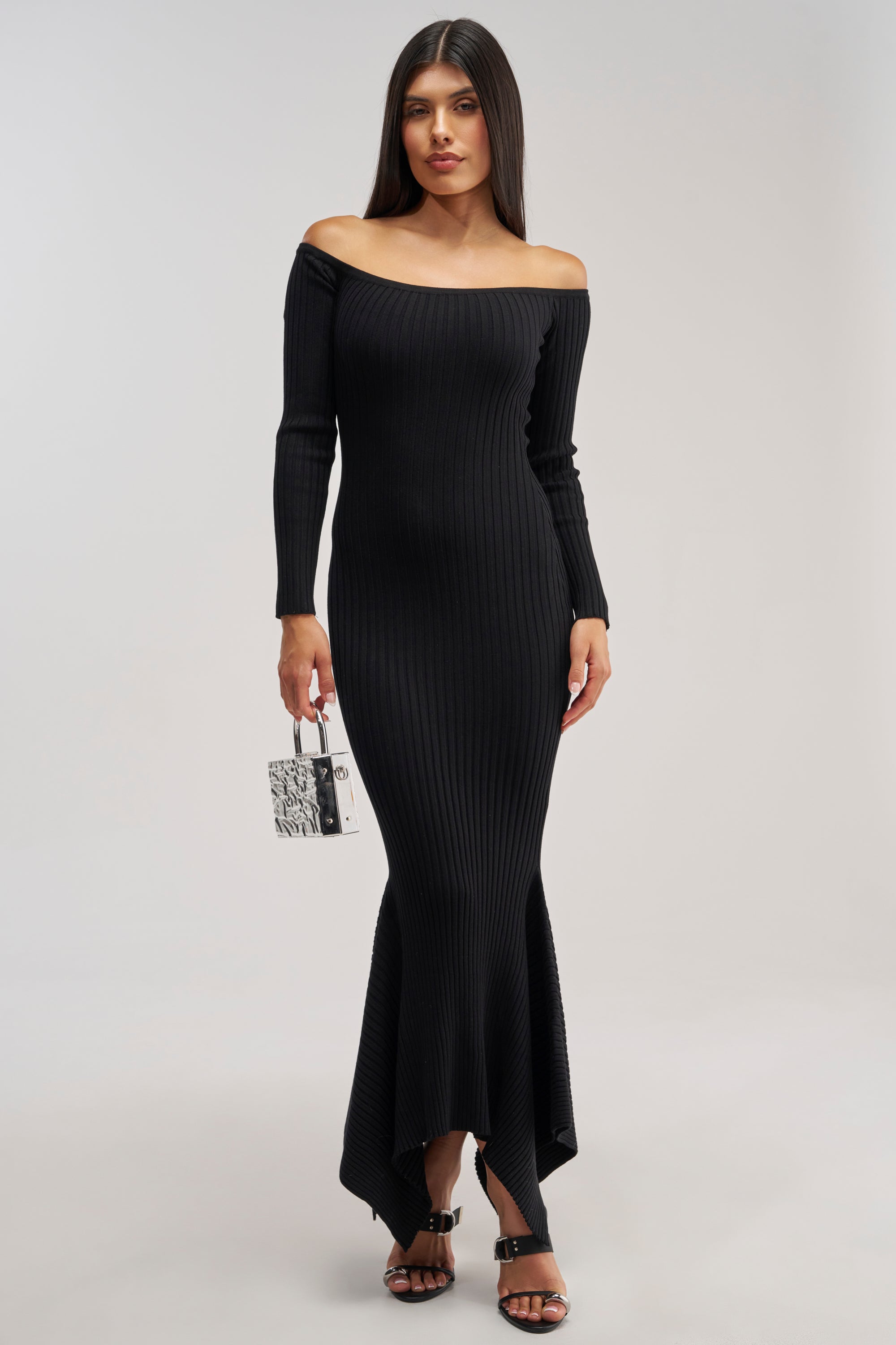 A woman wearing the SO SWEET KNIT MAXI DRESS IN BLACK—a long, off-the-shoulder sweater dress featuring ribbed detailing and long sleeves—pairs it with black heeled sandals and a small black-and-white handbag as she stands against a plain, light background. This chic dress is the perfect choice for a winter date night.