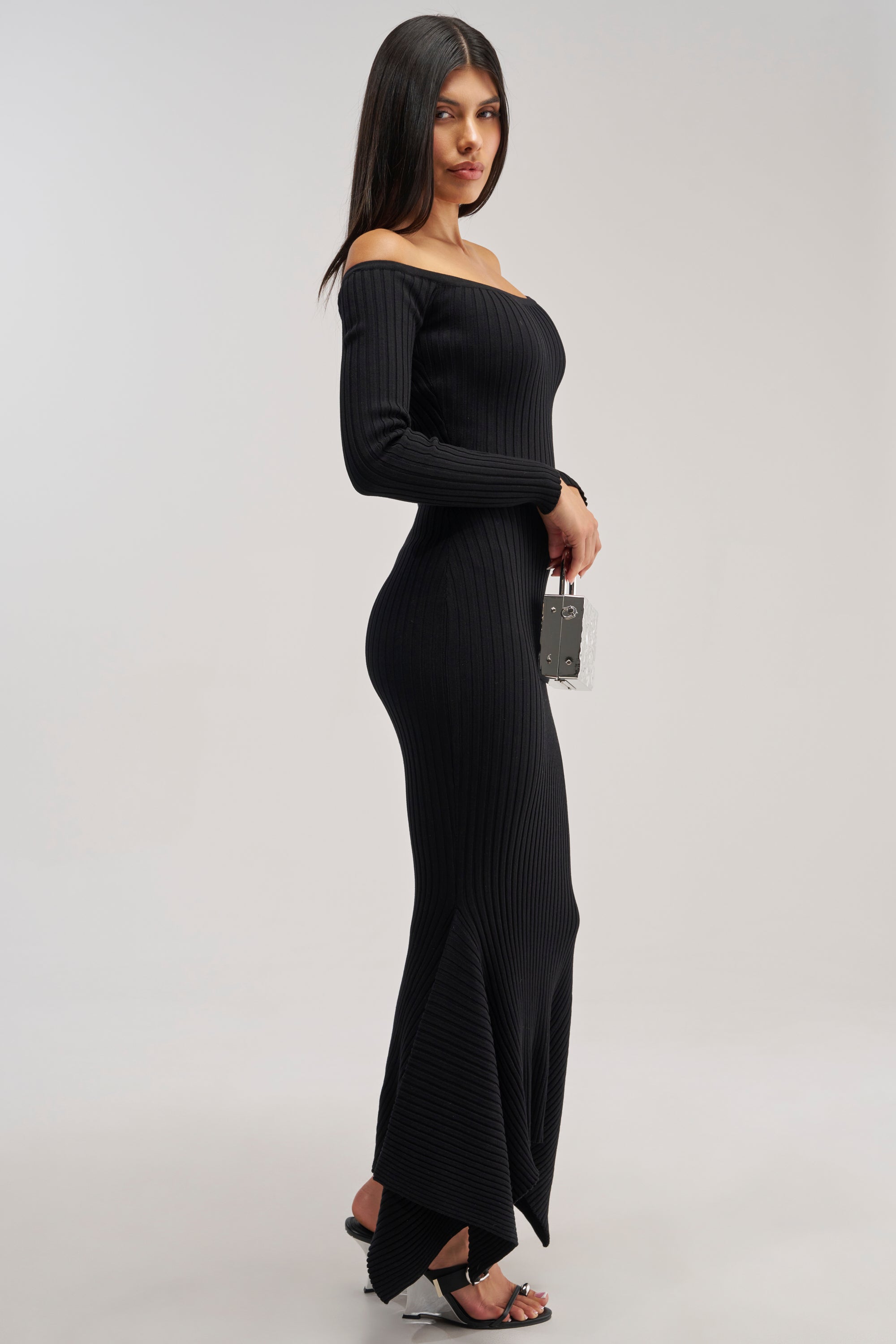 A woman stands sideways against a plain background, holding a small clear purse and wearing black heeled sandals. She is dressed in the SO SWEET KNIT MAXI DRESS IN BLACK—a ribbed, off-the-shoulder sweater dress that exudes effortless elegance and makes for an ideal winter date night choice.