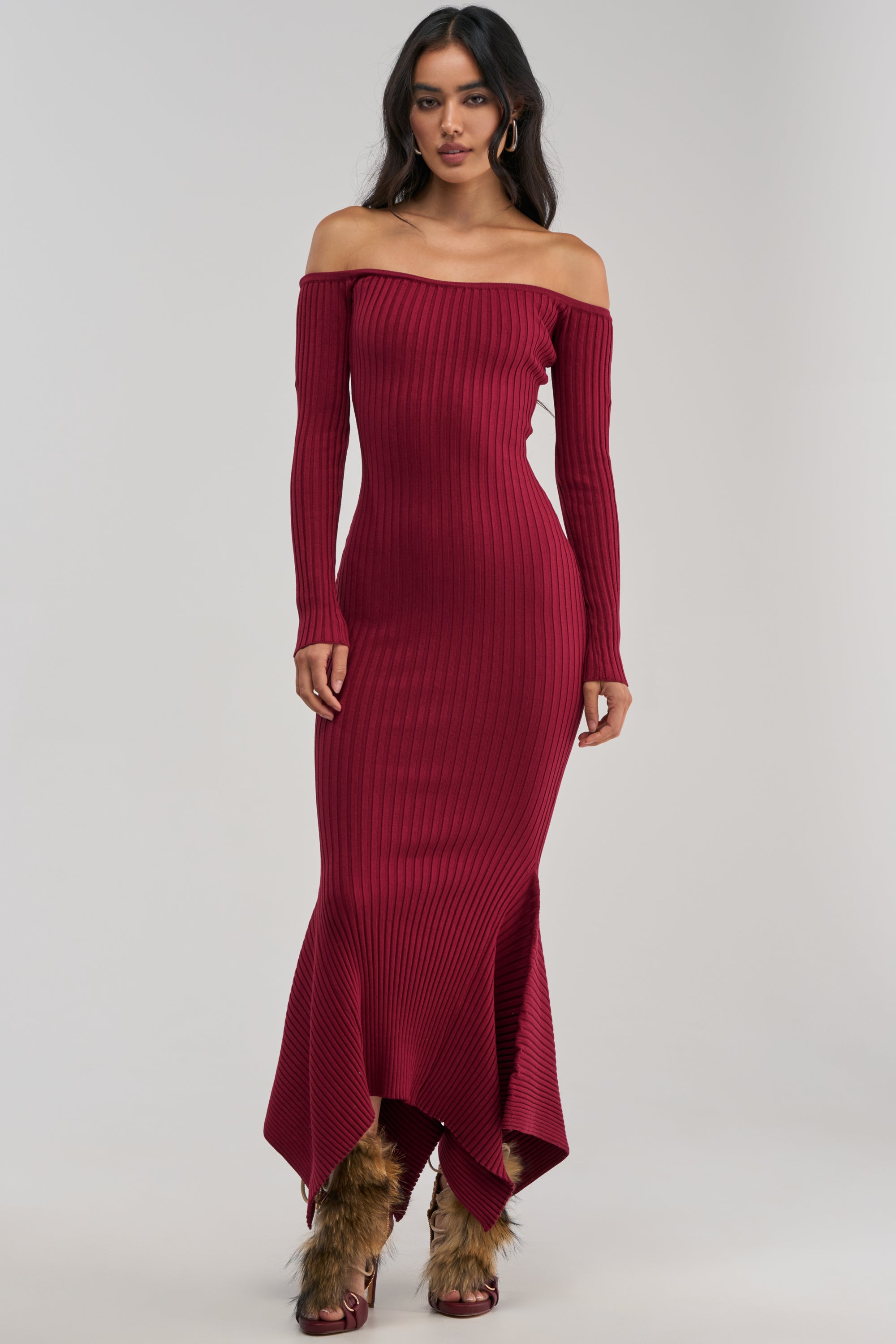 A woman stands against a plain light background wearing the SO SWEET KNIT MAXI DRESS IN BURGUNDY—an off-the-shoulder, long sleeve maxi dress with a fitted, ribbed texture and an asymmetrical hem—paired with fur-trimmed heeled sandals.