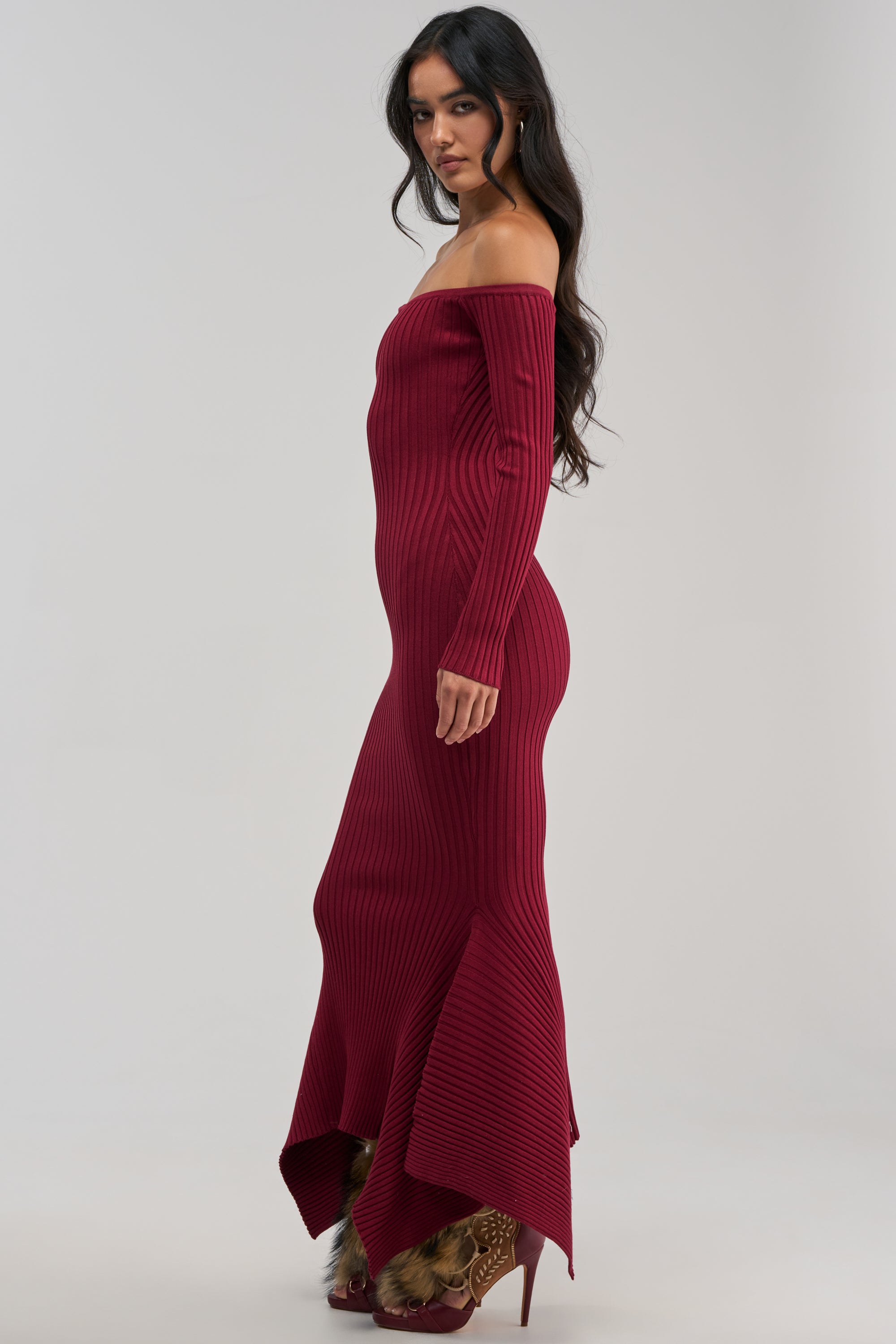 A woman stands sideways wearing the SO SWEET KNIT MAXI DRESS IN BURGUNDY, featuring a ribbed texture, off-shoulder neckline, and flared hem for a sleek mermaid silhouette. The rich burgundy hue beautifully complements her long, wavy dark hair as she gazes at the camera against a plain light background.