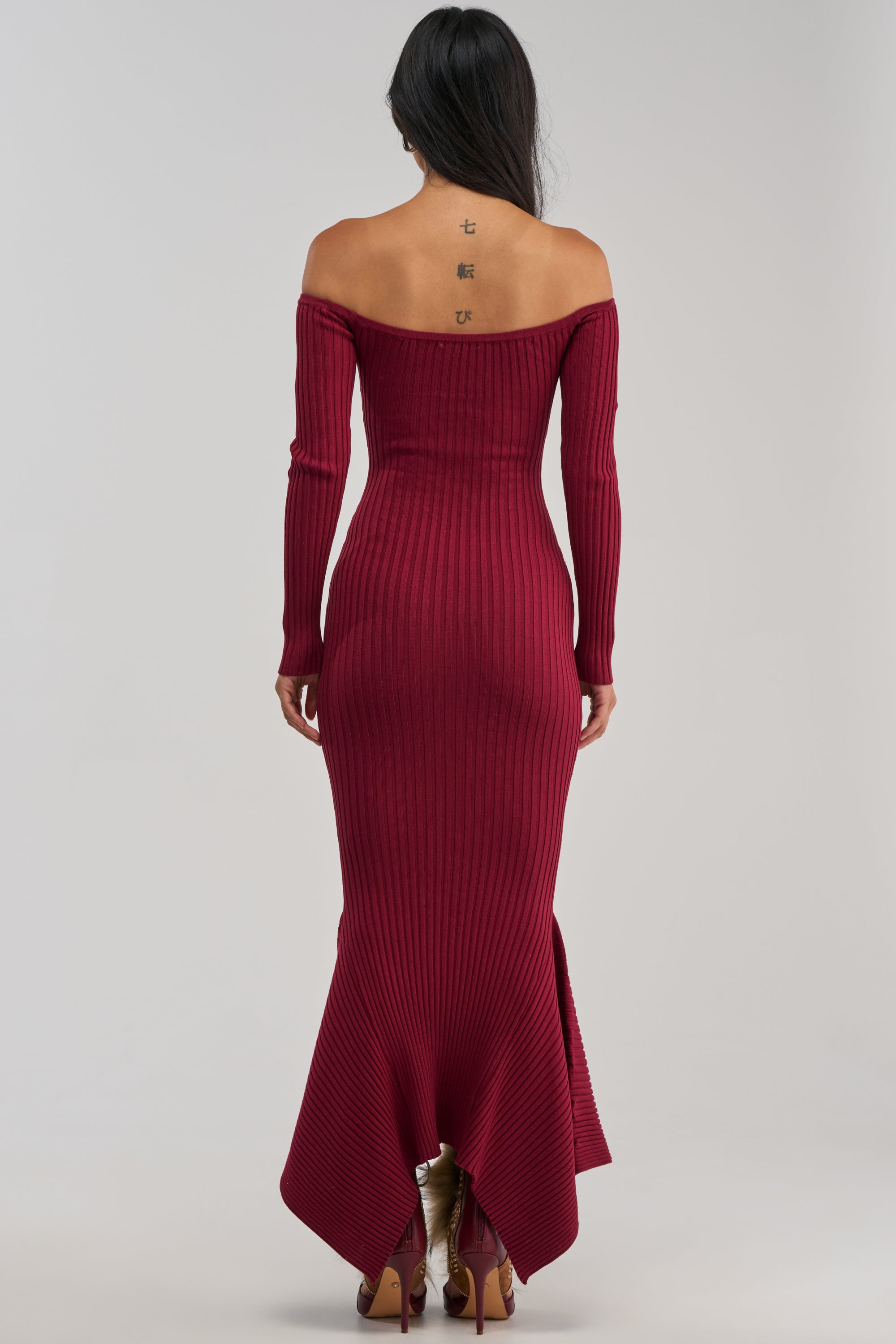 A woman stands with her back to the camera, wearing the SO SWEET KNIT MAXI DRESS IN BURGUNDY. The ribbed knit dress features an off-the-shoulder neckline, long sleeves, and a flattering mermaid silhouette that accentuates her figure. Three tattoos are visible on her upper back.