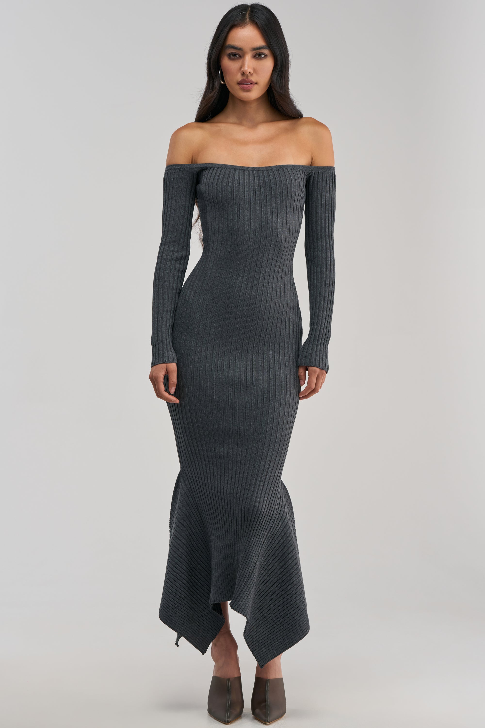 Wearing the SO SWEET KNIT MAXI DRESS IN HEATHER GREY—a long, dark gray off-the-shoulder ribbed knit dress featuring long sleeves and an asymmetrical hem that’s perfect for a winter date night—a woman stands against a plain light background.