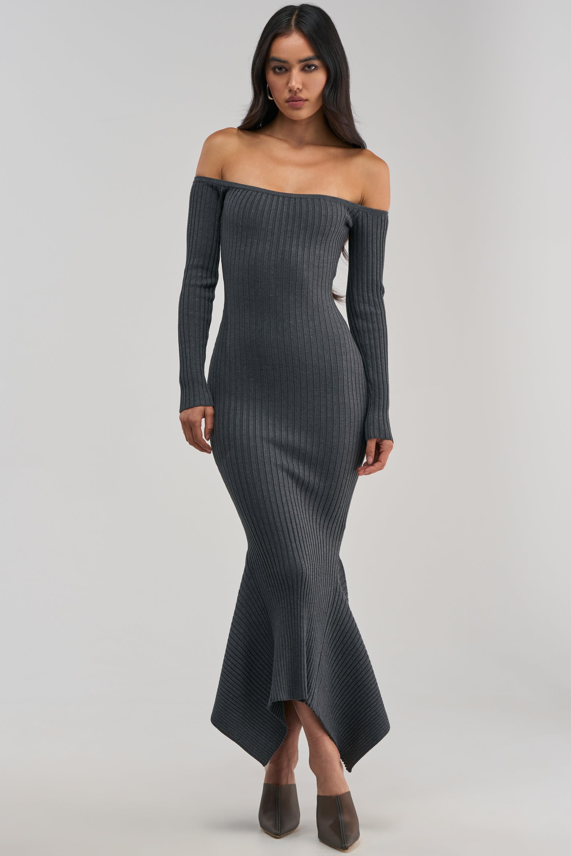 Dressed in the SO SWEET KNIT MAXI DRESS IN HEATHER GREY, this woman exudes effortless elegance as she stands against a plain light background. The ribbed sweater dress features a long, off-the-shoulder silhouette with fitted long sleeves and a flared hem, delivering a chic yet cozy vibe that’s perfect for a winter date night.