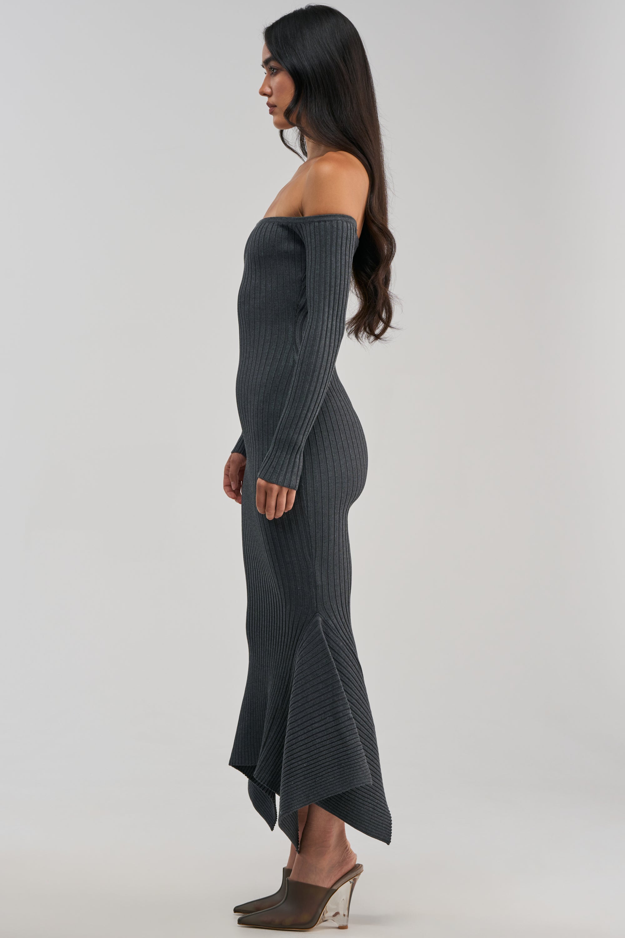 A woman stands sideways against a plain light background, wearing the SO SWEET KNIT MAXI DRESS IN HEATHER GREY—a long, ribbed maxi dress with an off-the-shoulder neckline, long sleeves, and an asymmetrical hem. Styled as the perfect winter date night outfit, she completes her look with taupe wedge heels.