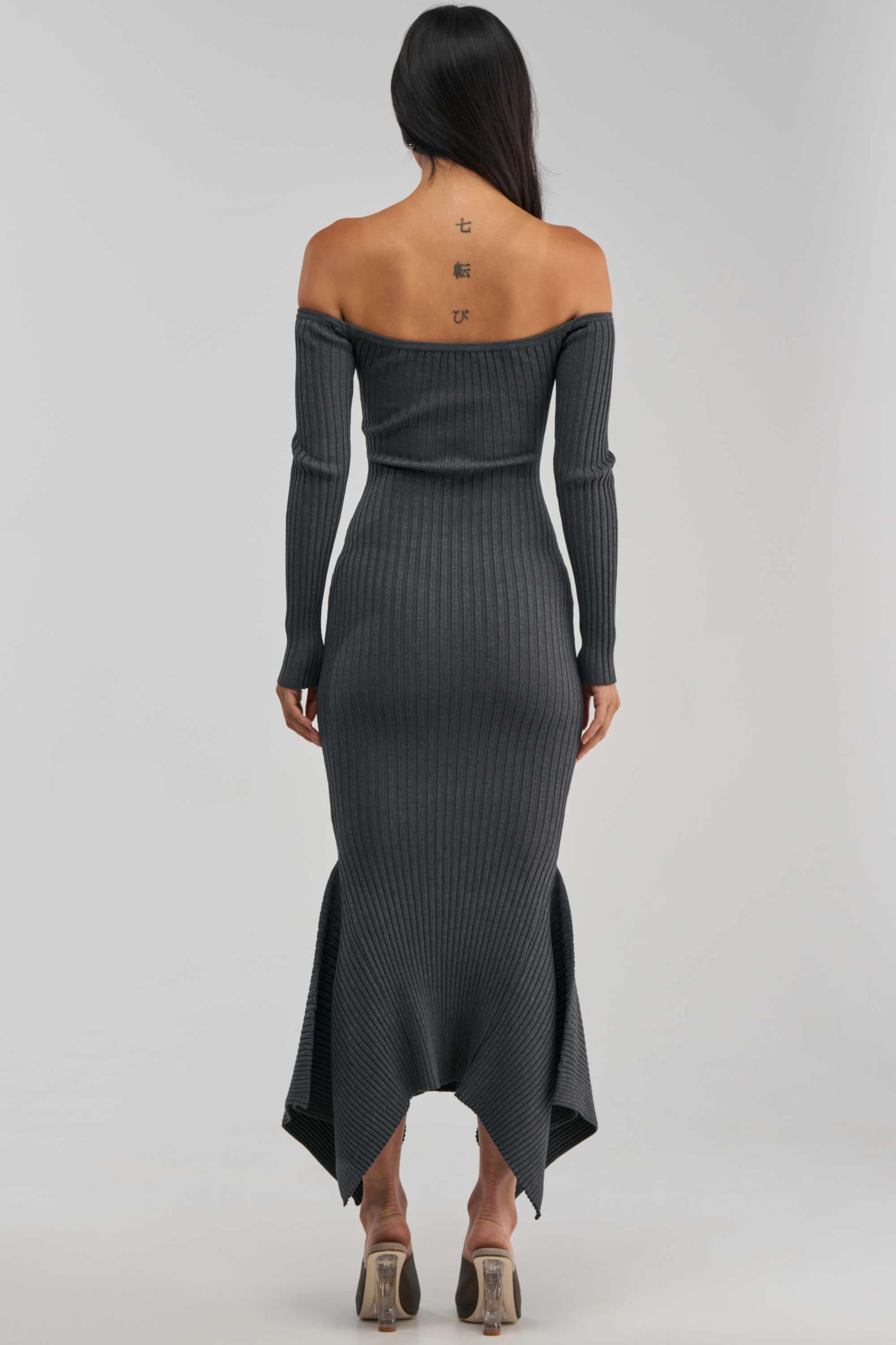 A woman with long dark hair stands with her back to the camera, showcasing three tattoos along her upper spine. She wears the SO SWEET KNIT MAXI DRESS IN HEATHER GREY, an off-the-shoulder ribbed sweater dress styled for a chic winter date night with clear-heeled shoes.