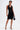 A woman stands confidently in the EDGY AND COOL LACE UP KNIT MINI DRESS, featuring black knit fabric, silver eyelets, and a bodycon fit. She holds a black handbag, wears heels, and shows arm tattoos against a plain white background.