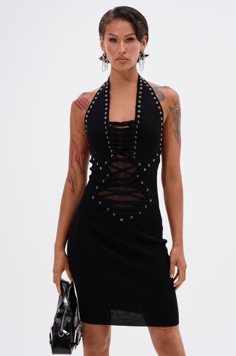A person with short hair and tattoos models the EDGY AND COOL LACE UP KNIT MINI DRESS, holding a black handbag against a plain white background.