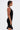 A woman with short dark hair stands in profile, wearing the Edgy and Cool Lace Up Knit Mini Dress—a black bodycon piece with silver studs—while holding a small black bag. She displays a large red abstract upper arm tattoo against a plain white background.