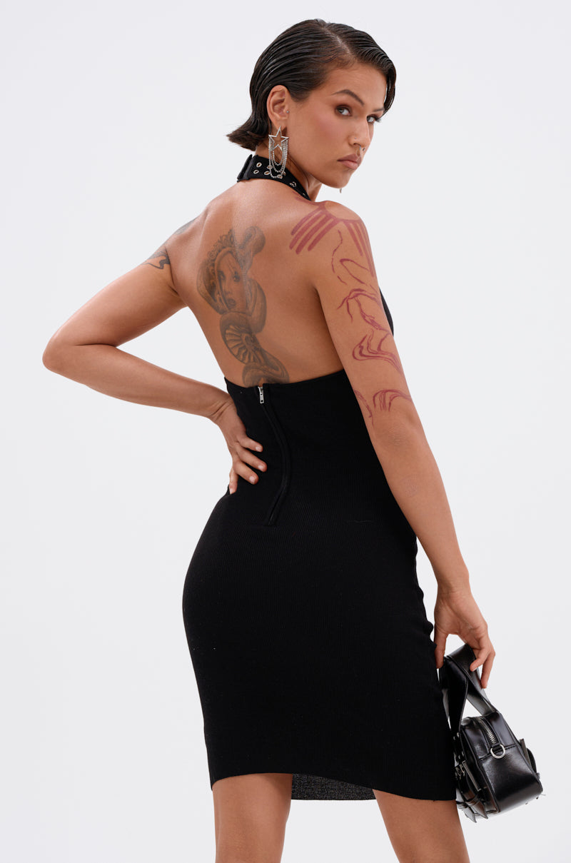 A confident individual with slicked-back hair, large earrings, and tattoos poses against a white background wearing the EDGY AND COOL LACE UP KNIT MINI DRESS—sleeveless, black, bodycon style—and holding a black handbag.