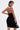 A confident individual with slicked-back hair, large earrings, and tattoos poses against a white background wearing the EDGY AND COOL LACE UP KNIT MINI DRESS—sleeveless, black, bodycon style—and holding a black handbag.