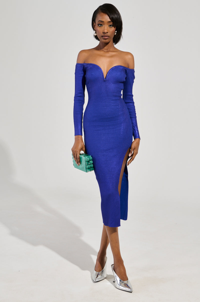 A person wearing the NOBODY'S BUSINESS BANDAGE MIDI DRESS IN BLUE, which features a curve-hugging fit, off-the-shoulder design, deep neckline, and high slit, is holding a small green clutch. They are standing against a neutral background and wearing silver pointed heels.