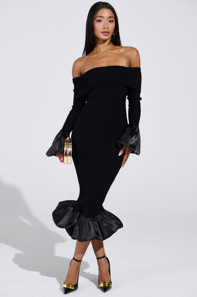 A woman poses in the BACK IN A MINUTE KNIT MIDI DRESS by AKIRA Label, featuring off-the-shoulder design, form-fitting silhouette, and ruffled satin cuffs and hem. She holds a gold clutch and pairs the dress with black and gold heels against a plain white background.