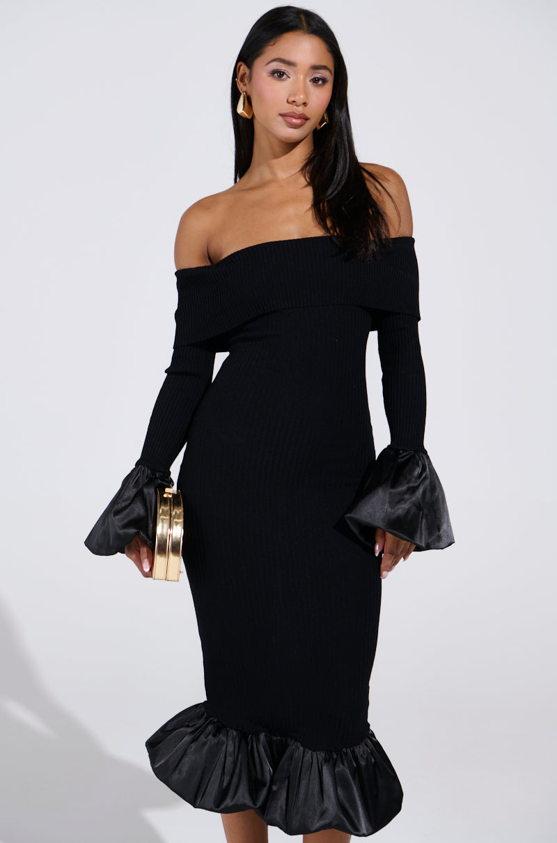 A woman in a BACK IN A MINUTE KNIT MIDI DRESS flaunts off-the-shoulder style with ruffled bell sleeves and hem, clutching a shiny gold cylindrical purse. Her long dark hair cascades elegantly against a plain white backdrop.
