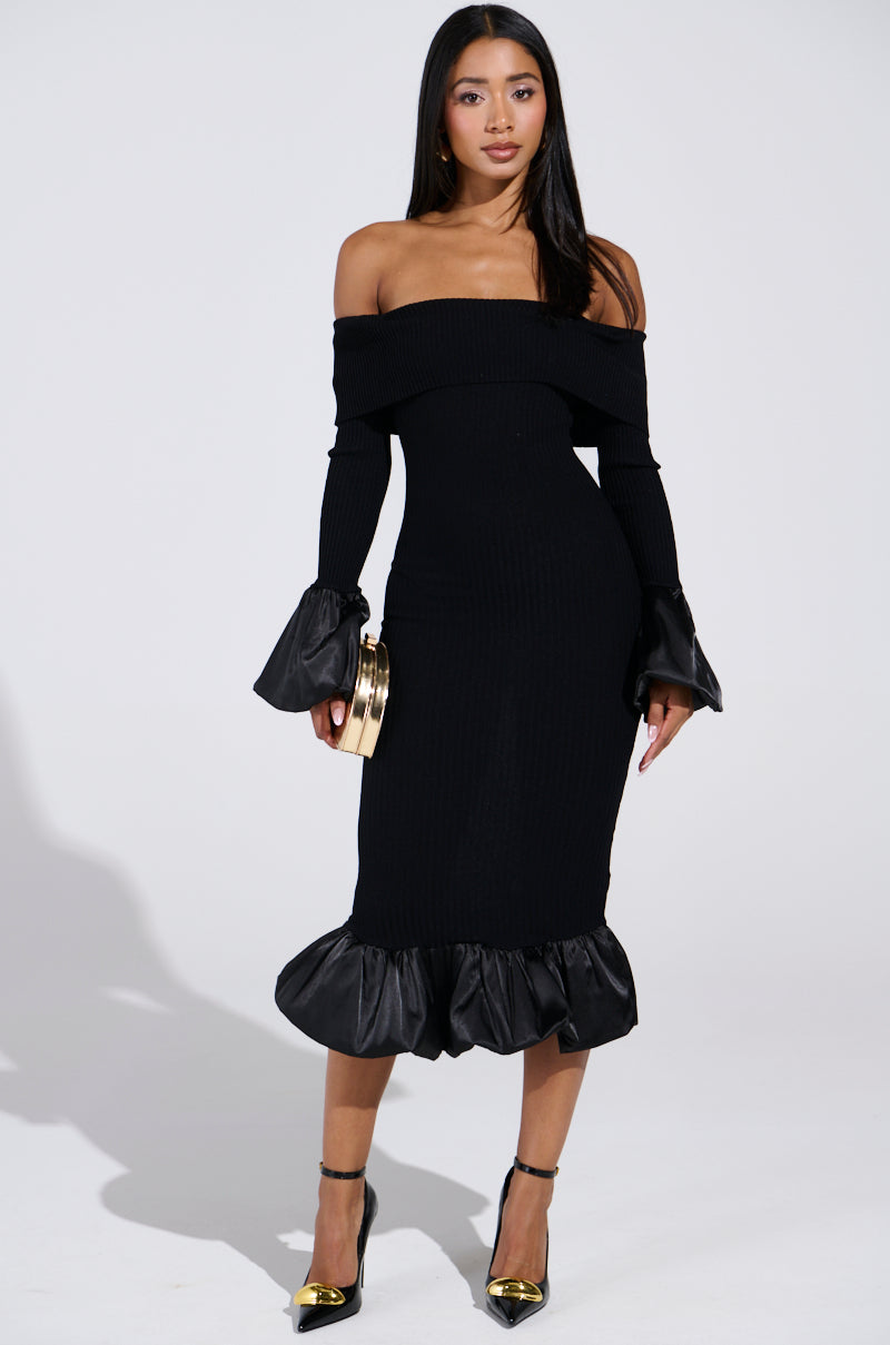 A woman confidently poses against a plain background in the BACK IN A MINUTE KNIT MIDI DRESS, featuring off-the-shoulder styling with flared sleeves and hem. She accessorizes with a small gold clutch and black and gold heels, her long hair cascading down as she gazes at the camera.