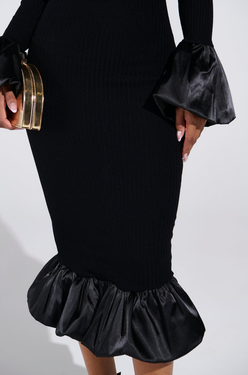 Wearing an AKIRA Label BACK IN A MINUTE KNIT MIDI DRESS with dramatic black, ruffled satin cuffs and hem, a person holds a shiny gold clutch in their left hand. The stunning off-the-shoulder design stands out against the plain, light-colored background.