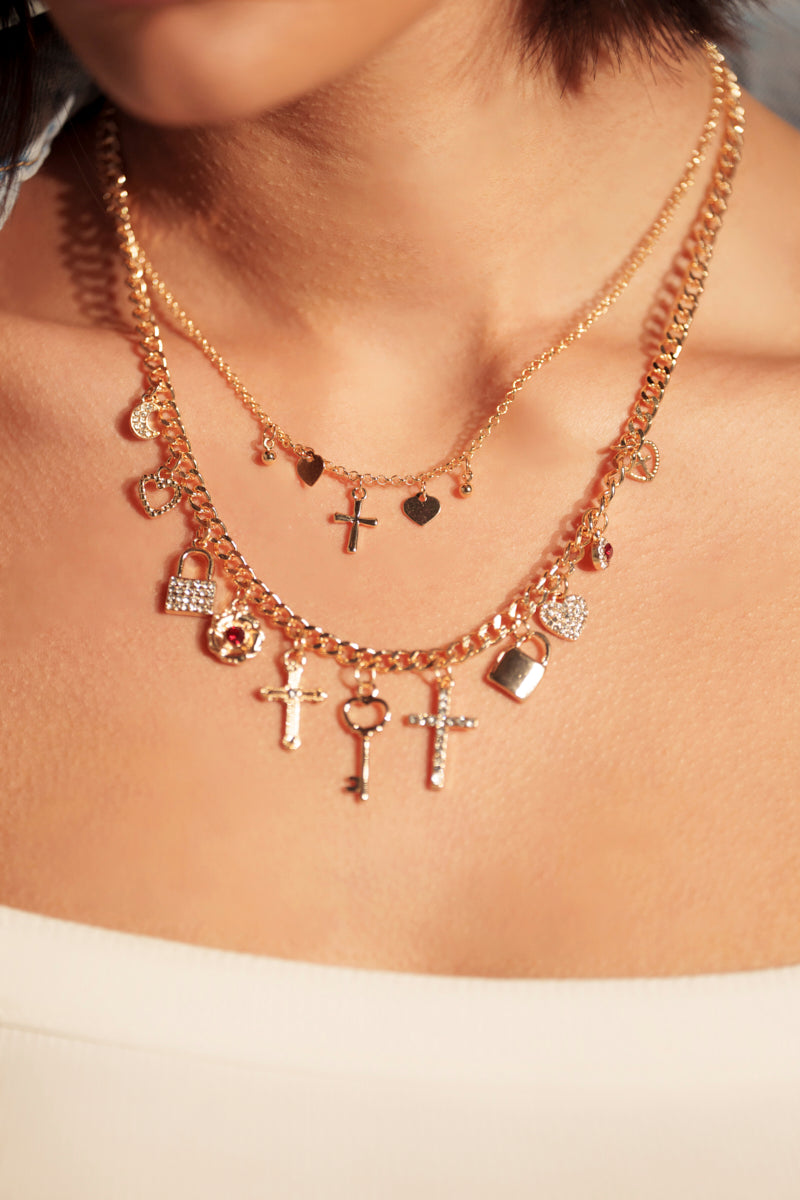 A close-up of a woman's neck wearing the CHARMED LIFE GOLD NECKLACE, a striking layered piece with two gold chains adorned with charms—crosses, hearts, keys, and padlocks—styled over a white top.