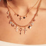 A close-up of a woman's neck wearing the CHARMED LIFE GOLD NECKLACE, a striking layered piece with two gold chains adorned with charms—crosses, hearts, keys, and padlocks—styled over a white top.