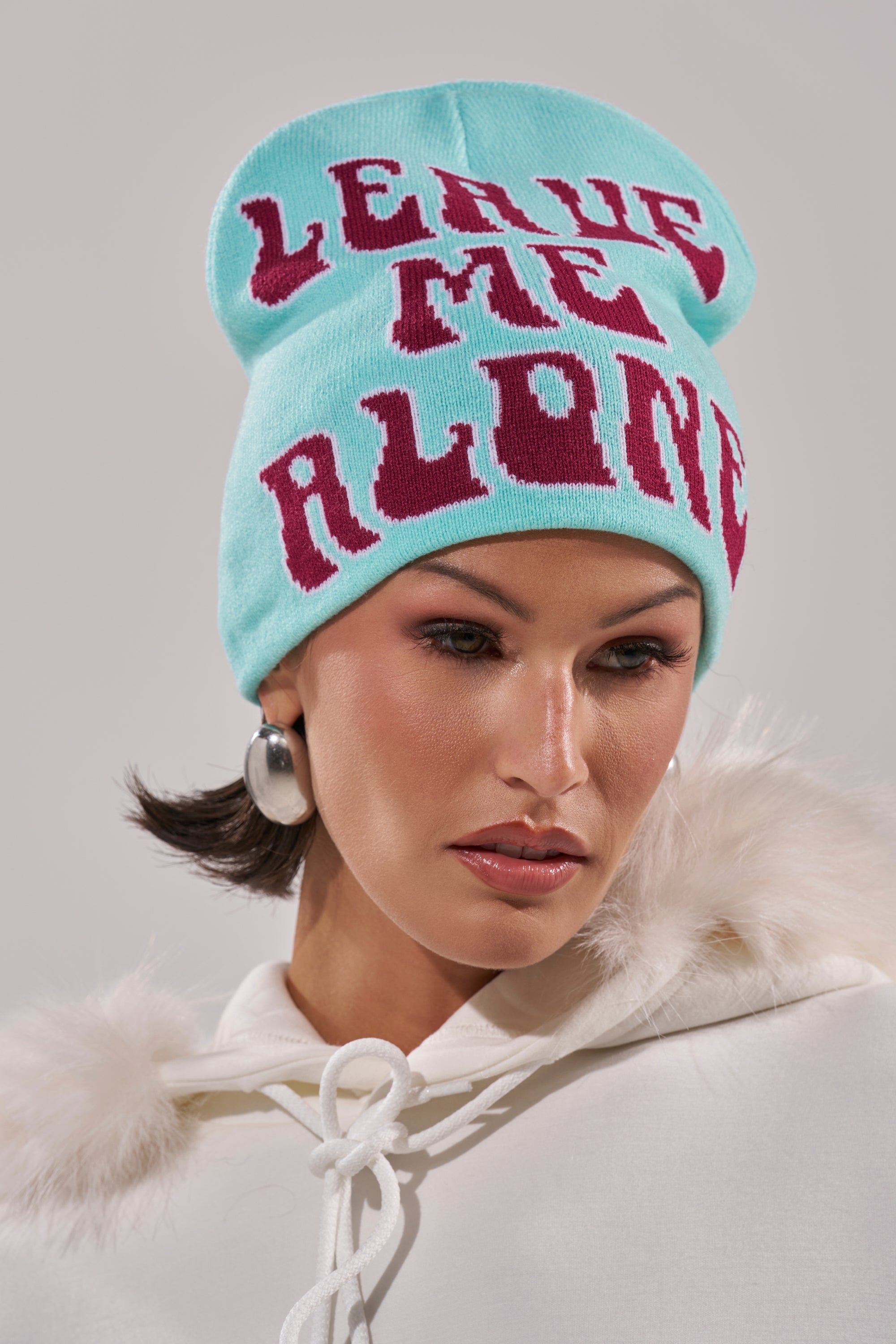 Wearing a light blue LEAVE ME ALONE BEANIE with bold pink letters, silver hoop earrings, and a white fur-trimmed hooded jacket, this person gazes slightly downward. 0