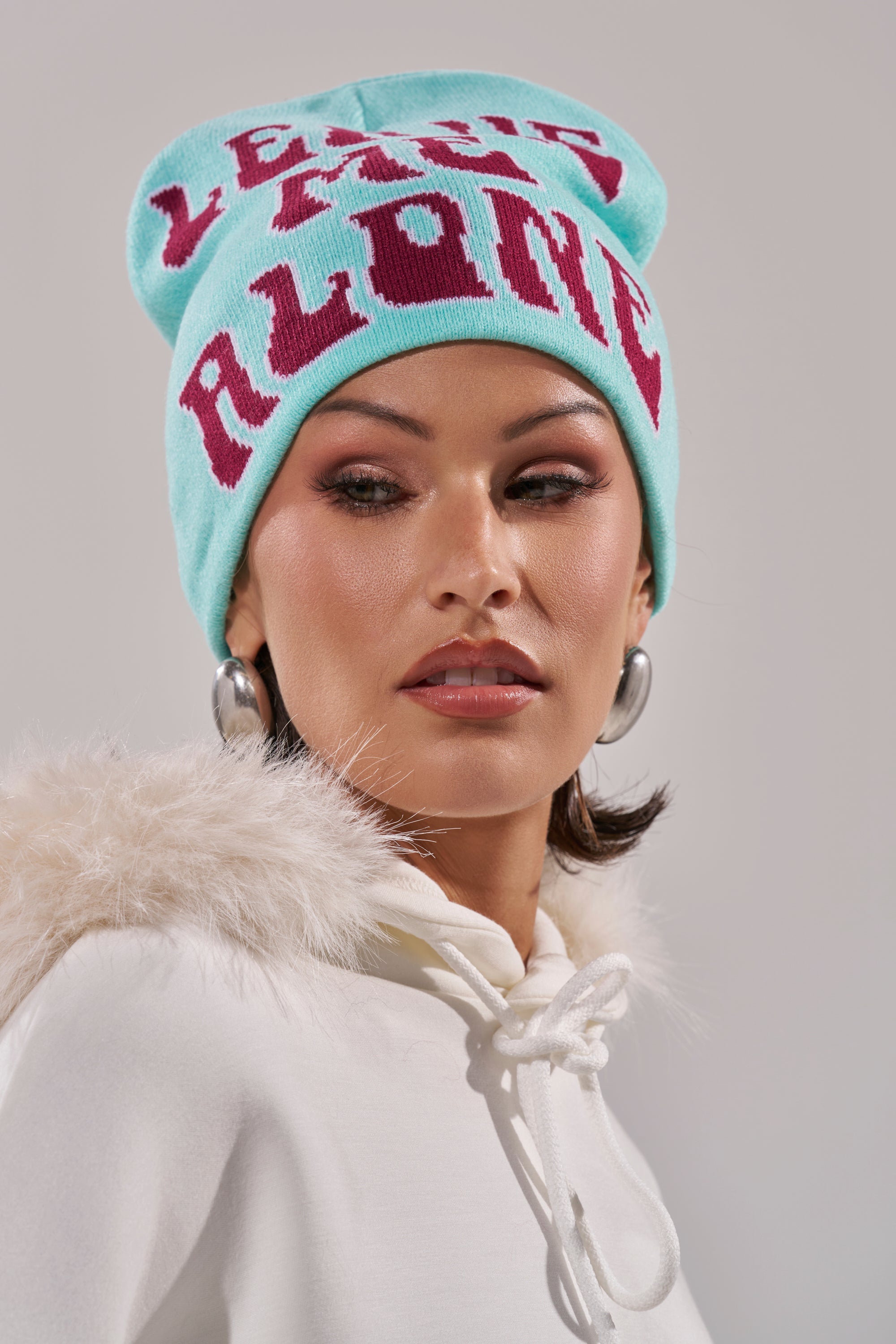 A woman wearing a light blue LEAVE ME ALONE BEANIE, a white furry-hooded sweatshirt, and large hoop earrings looks to the side against a plain background. 5