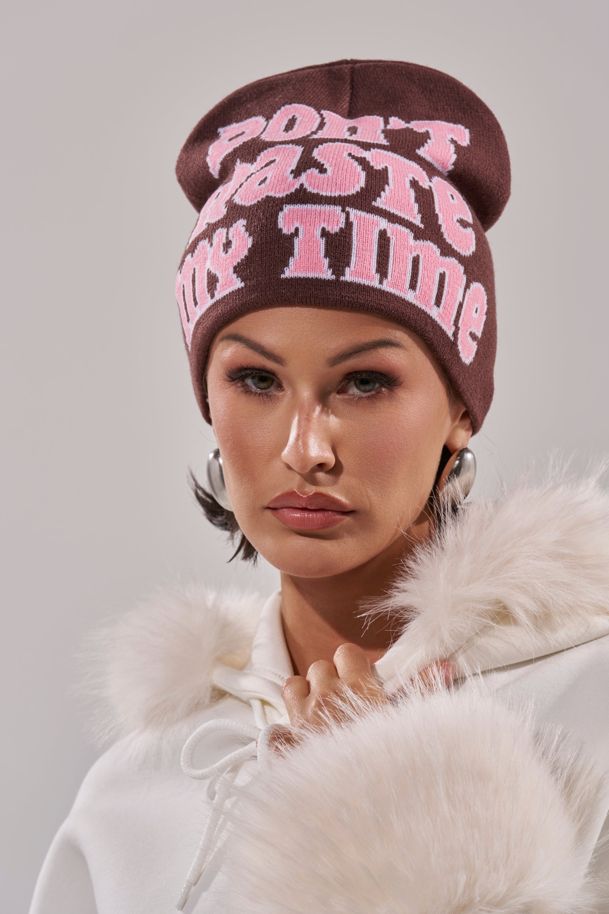 A woman wears the IM SO FORREAL BEANIE, a dark brown knit hat with bold pink "Don't Waste My Time" lettering, paired with a white fur-trimmed coat and large hoop earrings, gazing directly at the camera against a plain backdrop. 0