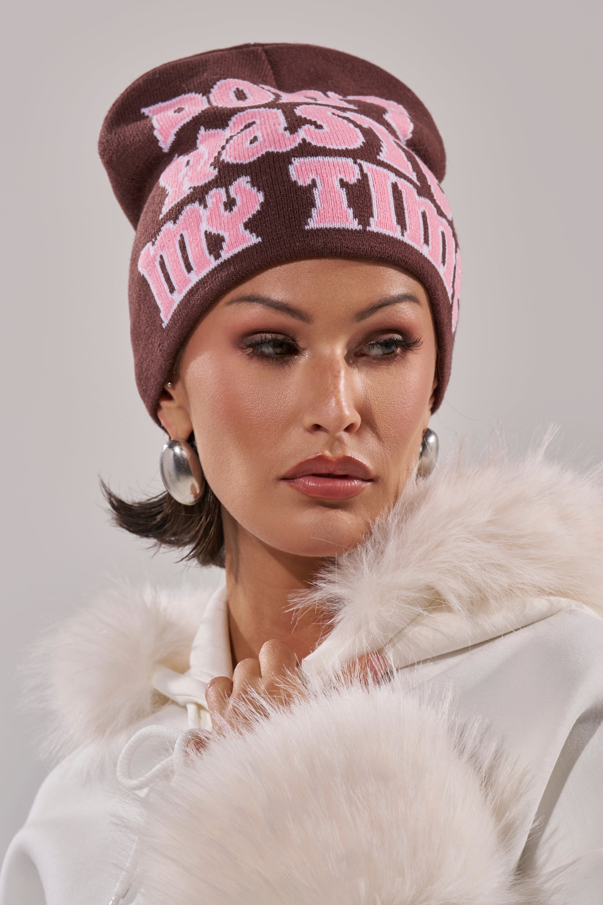 A woman with short dark hair, hoop earrings, and neutral makeup looks to the side wearing an IM SO FORREAL BEANIE in dark brown with pink text and a white faux fur coat. 5