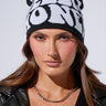 A woman with long brown hair is wearing the ALL WE NEED IS MONEY BEANIE, a black stretch knit hat featuring contrast text lettering. She completes her edgy look with a black leather jacket and a silver chain necklace, all set against a neutral background.