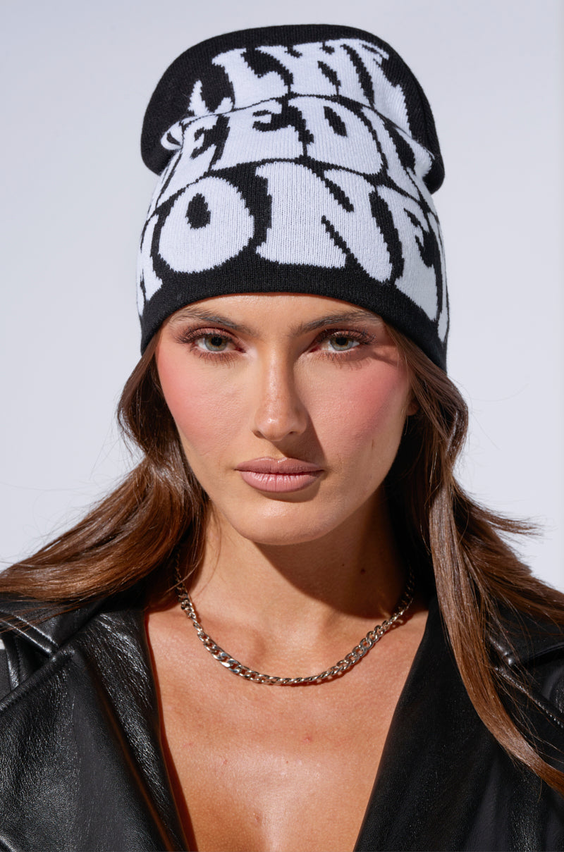 Wearing an ALL WE NEED IS MONEY black beanie, featuring a bold white text design, a person with long brown hair and a black leather jacket gazes directly at the camera against a plain light-colored background. 0