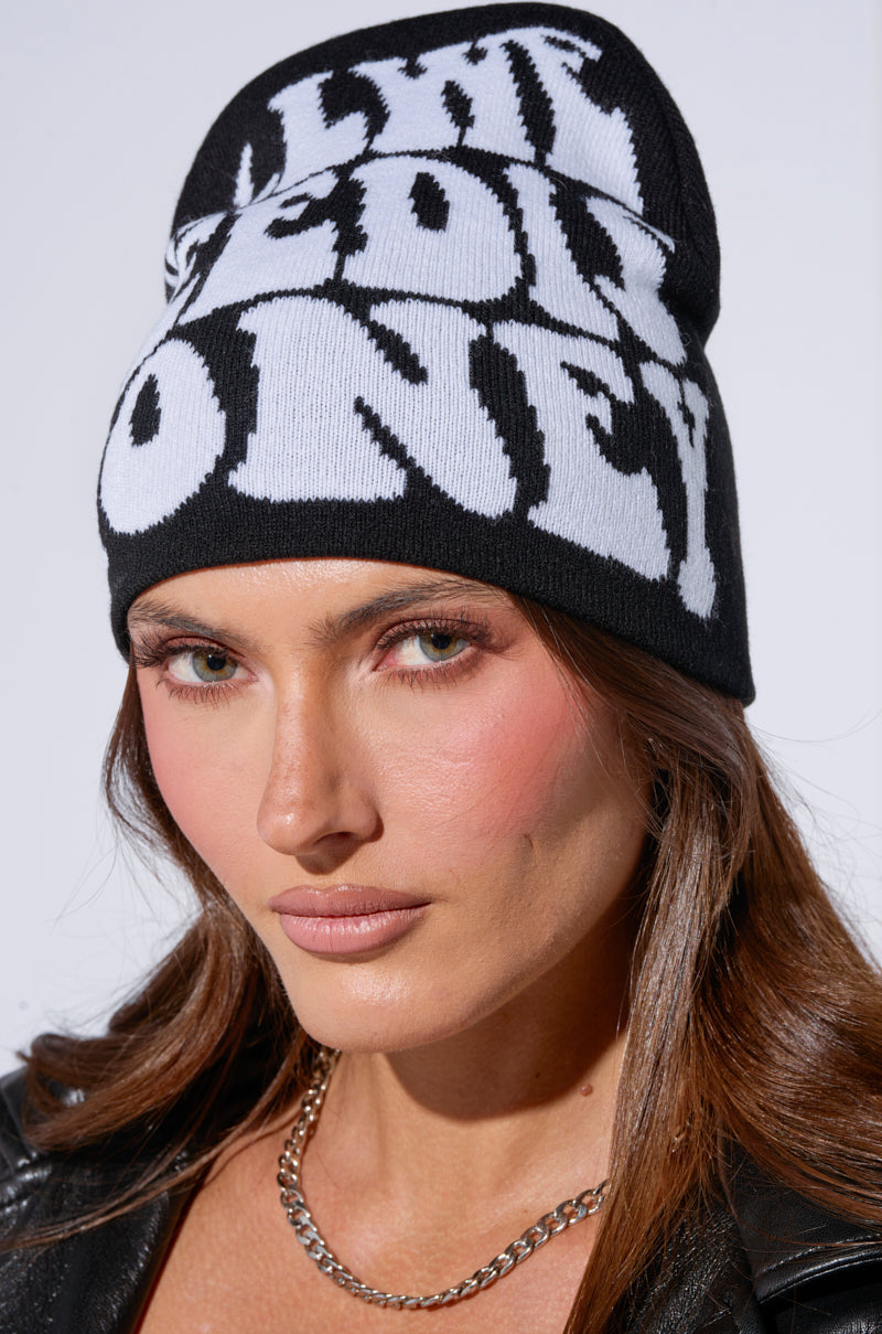 A person wears the "ALL WE NEED IS MONEY" black beanie and a leather jacket against a plain, light-colored wall. 1
