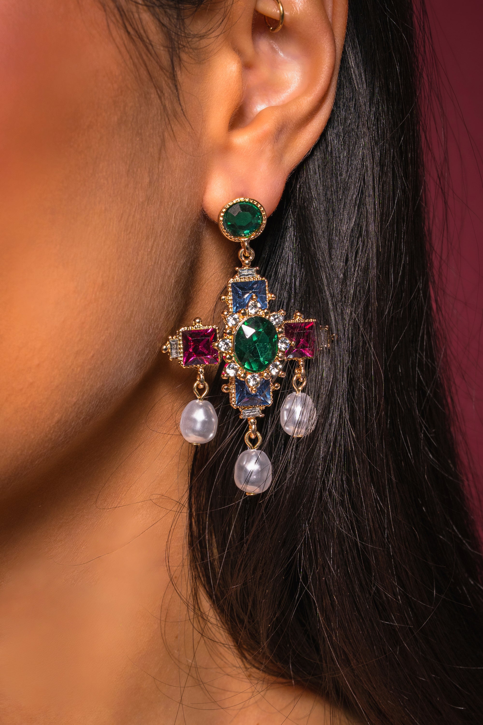 A close-up of a woman's ear wearing the GLAM EARRING, which features multi-color crystal gems, pearl-like dangles, and gold detailing, set against a dark background. 0