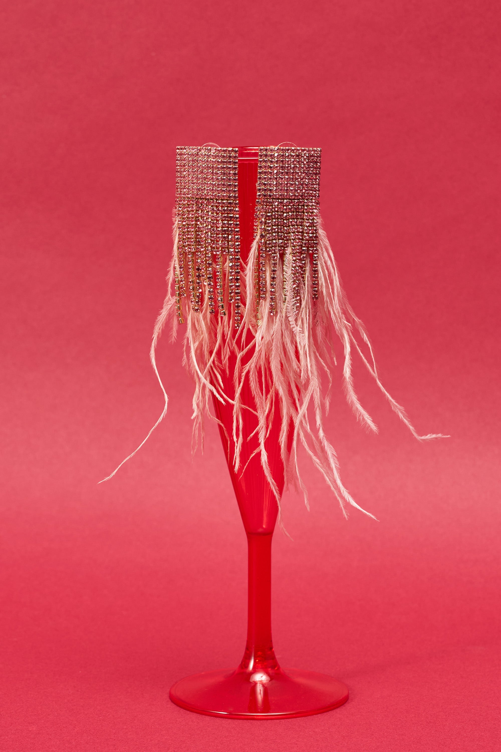 The SHAKE IT EARRING, with its beaded fringe and long, white feathery strands, is displayed draped over the rim of a red champagne flute against a matching red background, evoking elegant feather earrings. 0