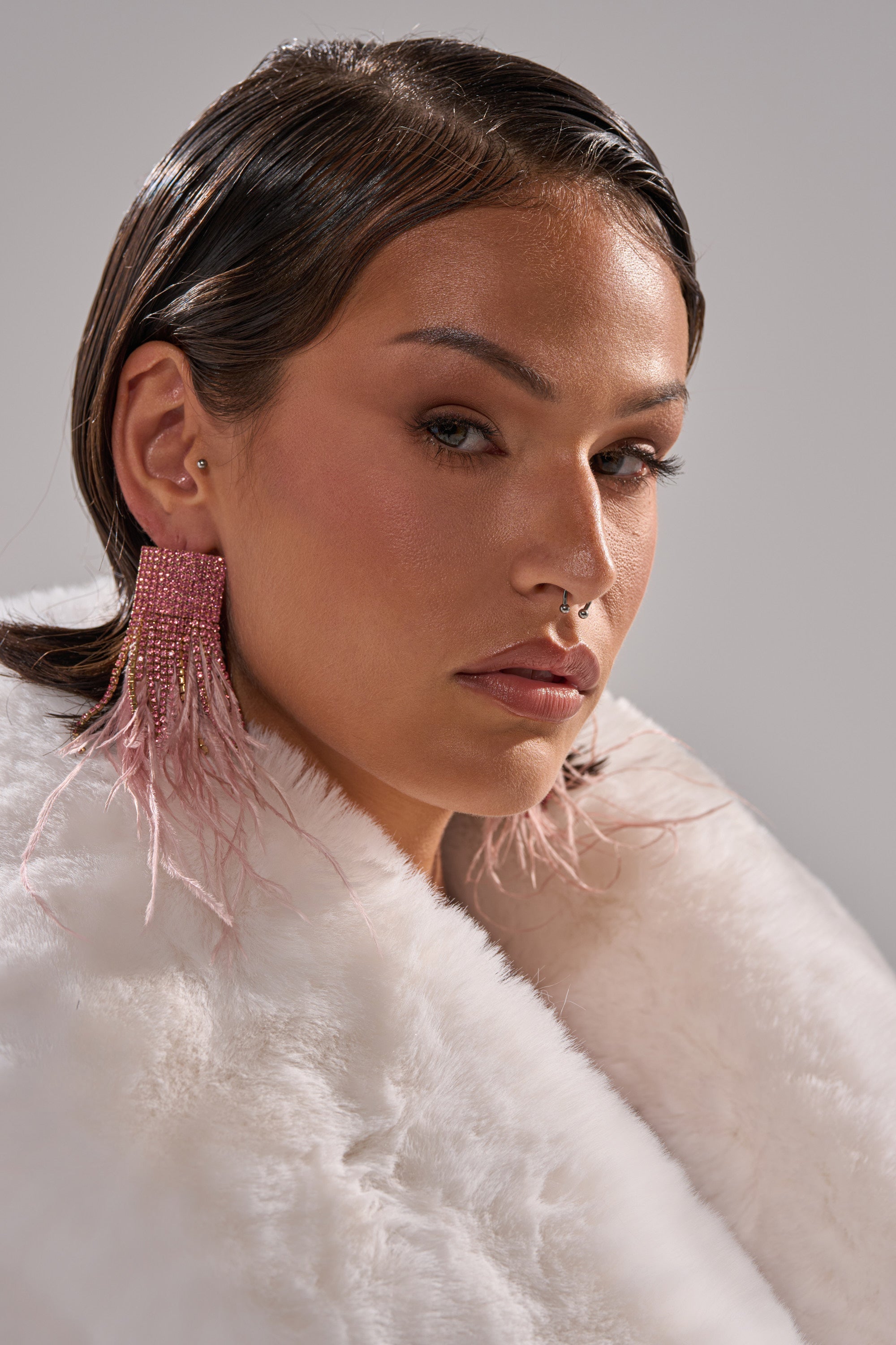 Wearing the SHAKE IT EARRING, a person in a white faux fur coat looks confidently at the camera. Their slicked-back hair, nose ring, and natural makeup complete the bold look. 3