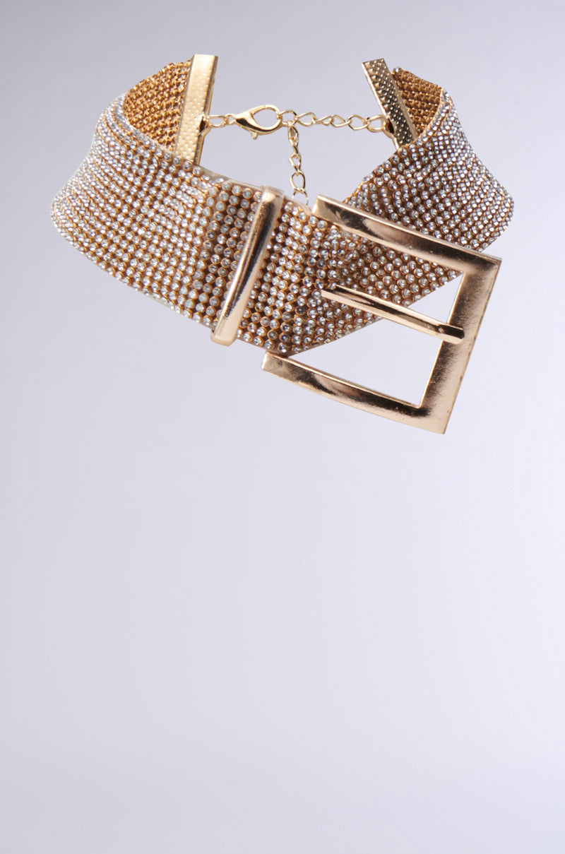 The BUCKLE UP BLING CHOKER, featuring a shimmering gold mesh design, boasts a metallic buckle pendant and rhinestone chain. It comes with an adjustable clasp, showcased against a gradient gray background.