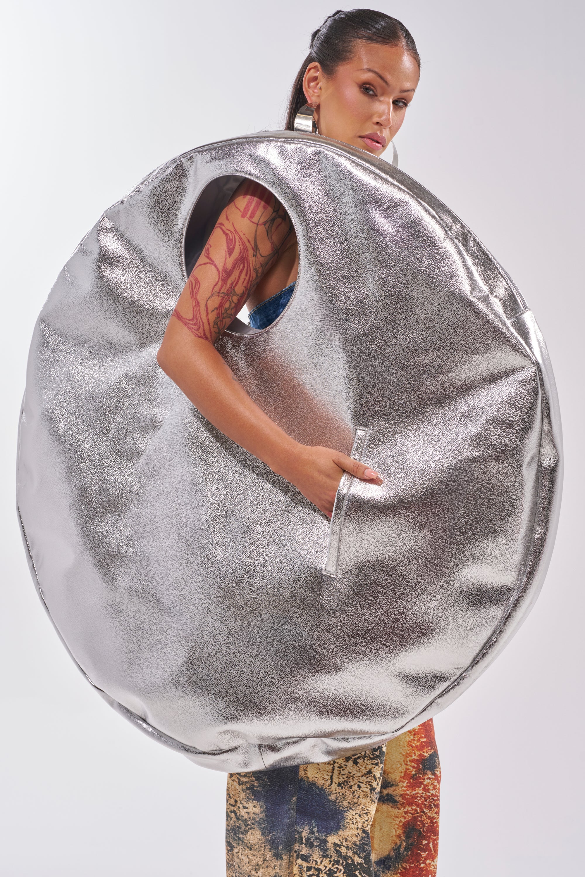 A woman models the OVERSIZED METAL MANIA PURSE—a large, round, metallic silver accessory with an armhole. She has long dark hair, bold earrings, a visible arm tattoo, and wears colorful patterned pants against a white backdrop. 0