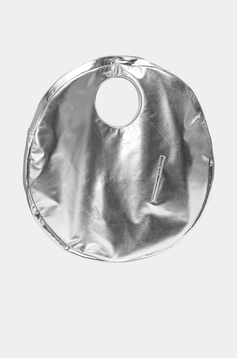 The OVERSIZED METAL MANIA PURSE is a round, oversized bag in silver faux leather with a cut-out handle and small diagonal front zipper pocket, shown against a plain light gray backdrop—a statement high fashion accessory. 1