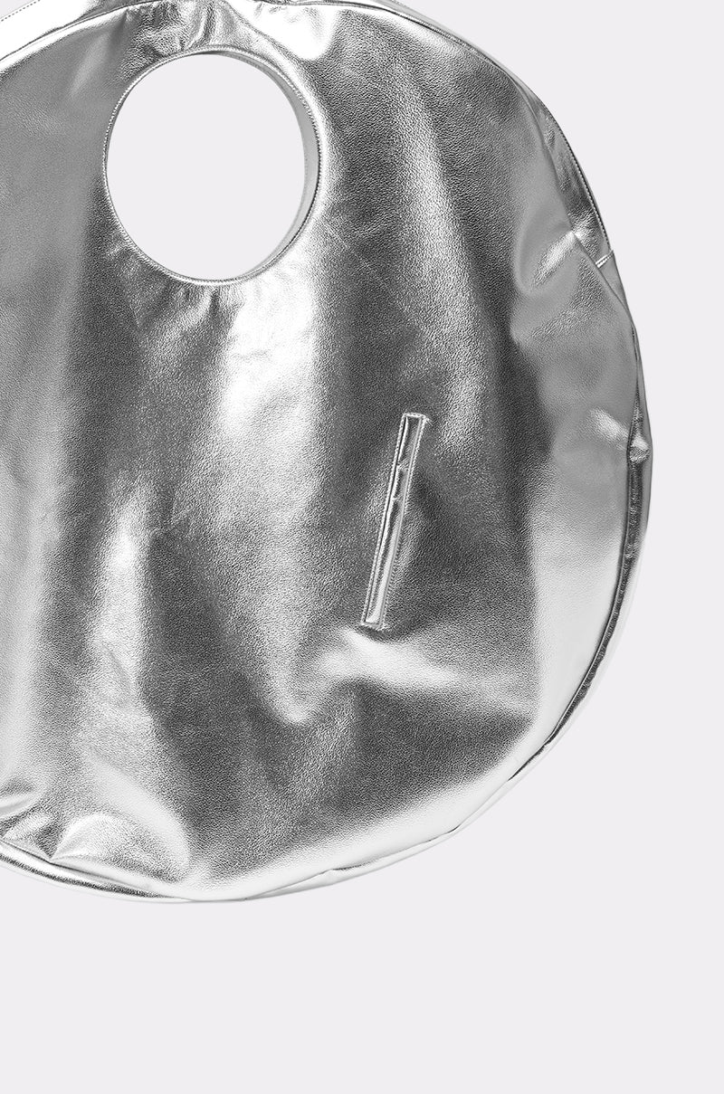The OVERSIZED METAL MANIA PURSE is a high-fashion oversized circular tote in metallic silver faux leather, with a round handle cutout and front zippered pocket, shown against a plain light background. 2