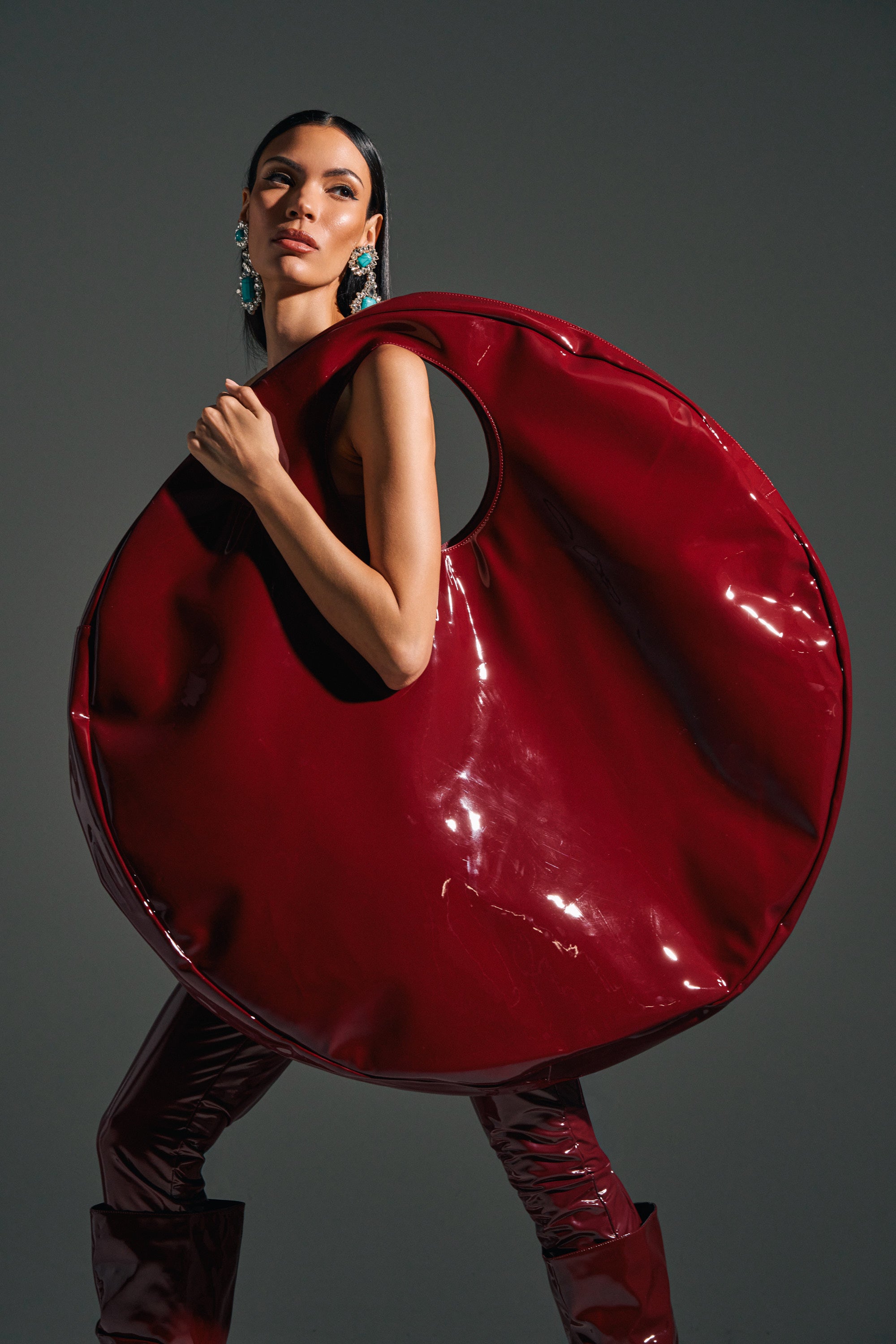A woman poses with the EPITOME OF LUXURY PATENT OVERSIZED TOTE, a glossy red faux patent leather bag featuring an oversized circular design and arm cut-out. She wears statement earrings, her hair sleek, against a dark gray background.
