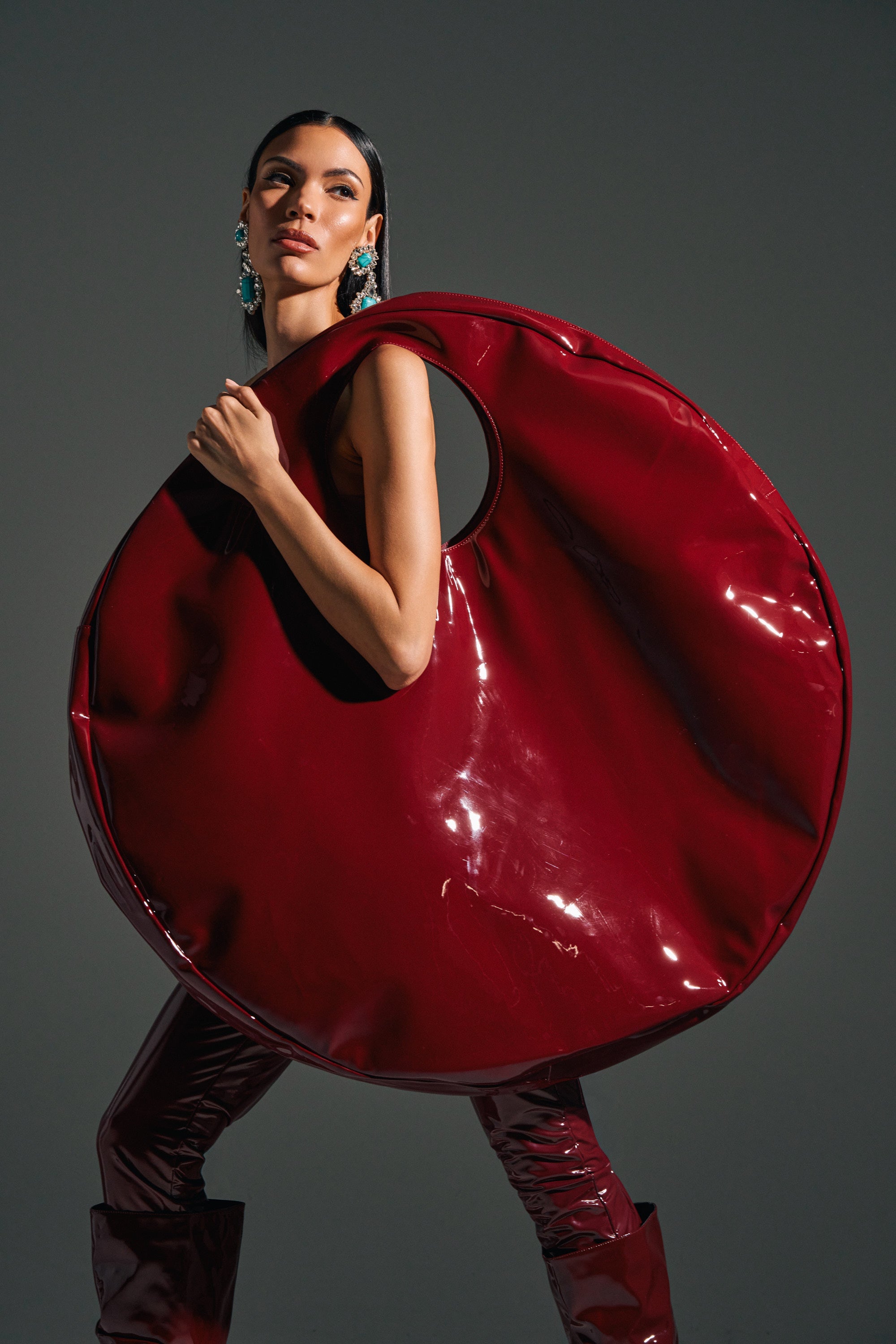 A woman poses with the EPITOME OF LUXURY PATENT OVERSIZED TOTE, a glossy red faux patent leather bag featuring an oversized circular design and arm cut-out. She wears statement earrings, her hair sleek, against a dark gray background. 0