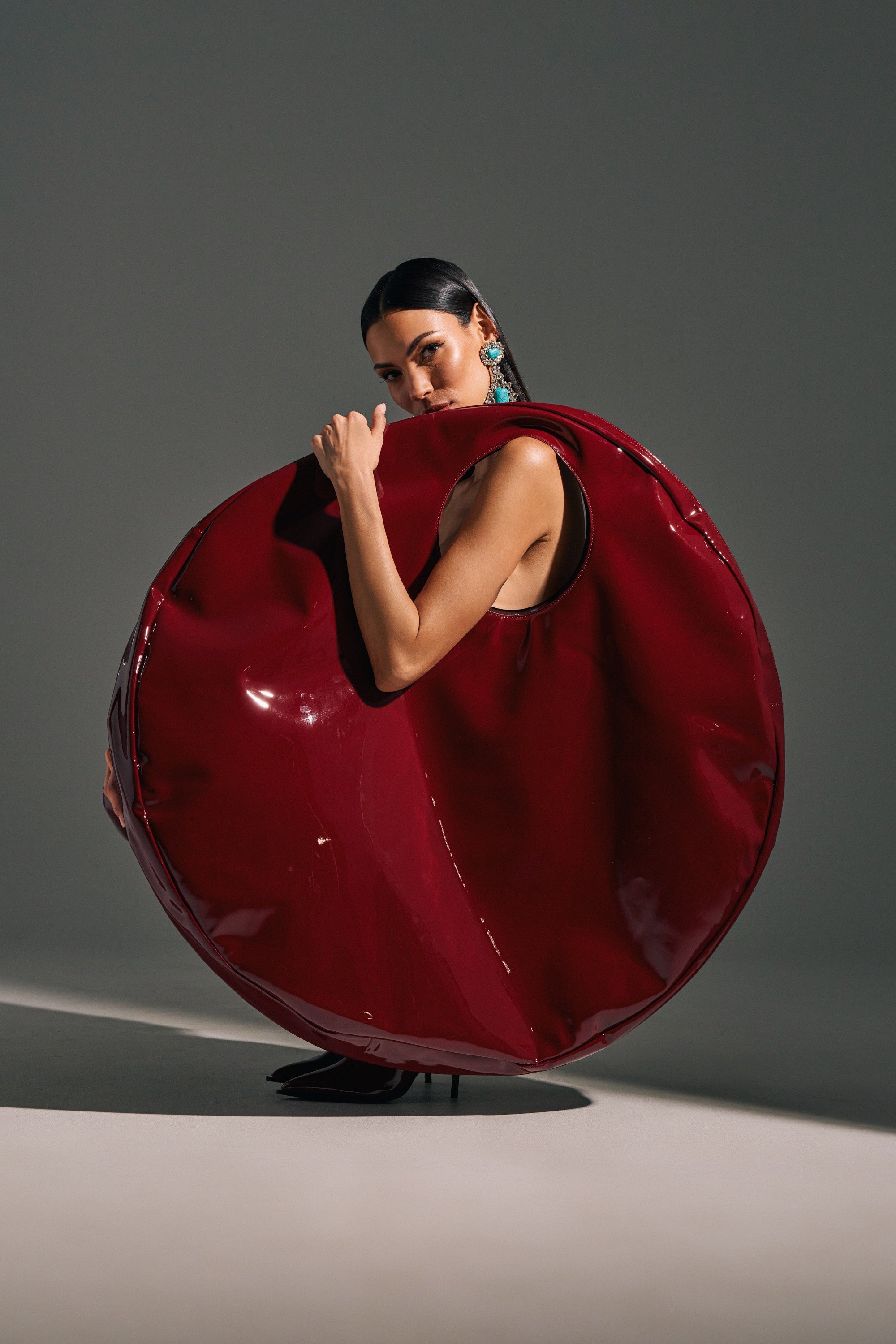 A woman poses with the EPITOME OF LUXURY PATENT OVERSIZED TOTE, a shiny deep red faux patent leather accessory. She pairs it with statement earrings and black heels, standing against a simple neutral background. 1