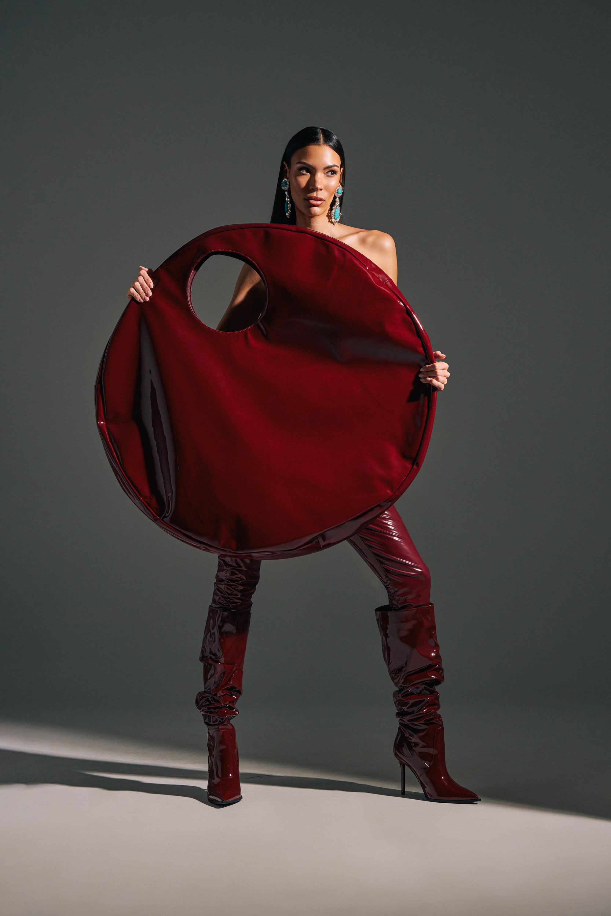 A woman in glossy burgundy boots holds the EPITOME OF LUXURY PATENT OVERSIZED TOTE, featuring matching faux patent leather and a cutout handle. She wears statement earrings, has straight dark hair, and stands confidently against a plain gray background.