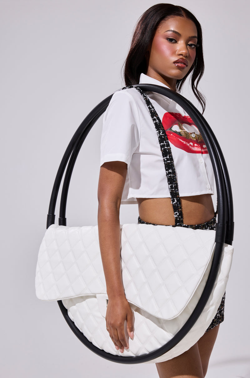A woman sporting a cropped white shirt with a lip graphic poses inside the IYKYK OVERSIZED PURSE, an oversized quilted faux leather bag featuring large black handles, set against a plain light background. 0