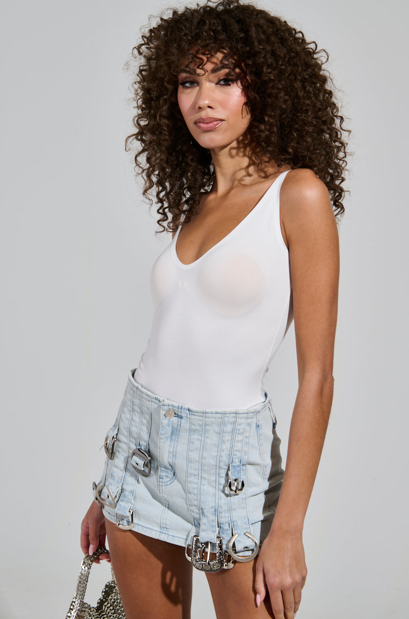 A person with curly hair poses confidently in a figure-flattering fit, showcasing the TIME TO GO BODYSUIT IN WHITE paired with light blue denim shorts adorned with buckles. They are holding a small textured handbag, standing against a plain background.