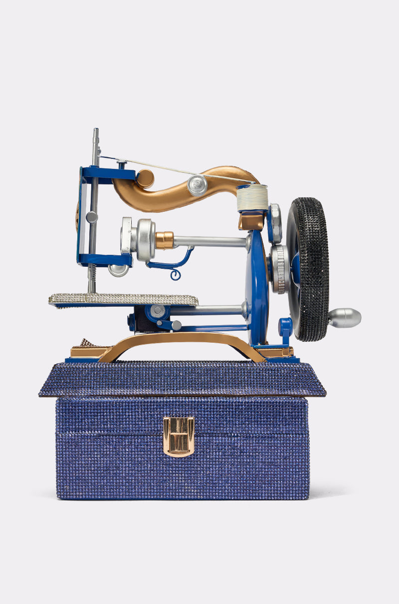 The SEW IT BAG, a vintage sewing machine-inspired handbag in blue and gold, rests on a matching textured blue box with a gold clasp, creating a charming display against a plain white background. 0